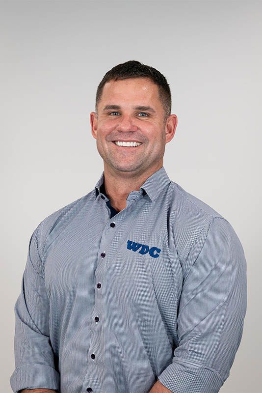 Smiling man in a gray button-down shirt with a blue logo, standing against a plain light background.