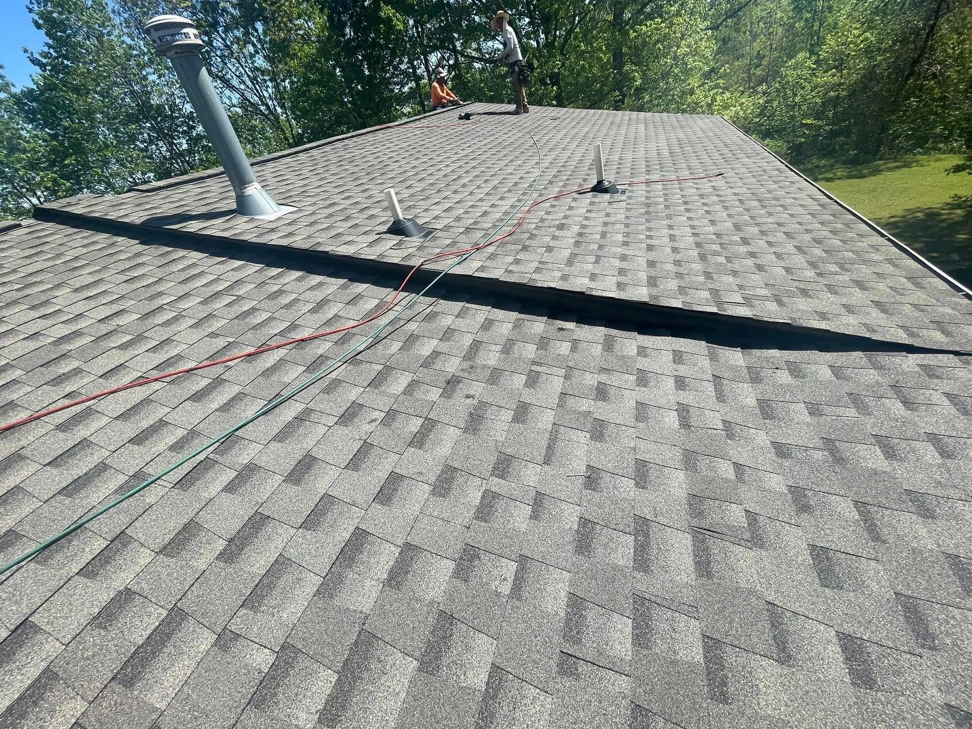 Gray shingle roof with vent pipes and sunlight, viewed from above, with trees in the background