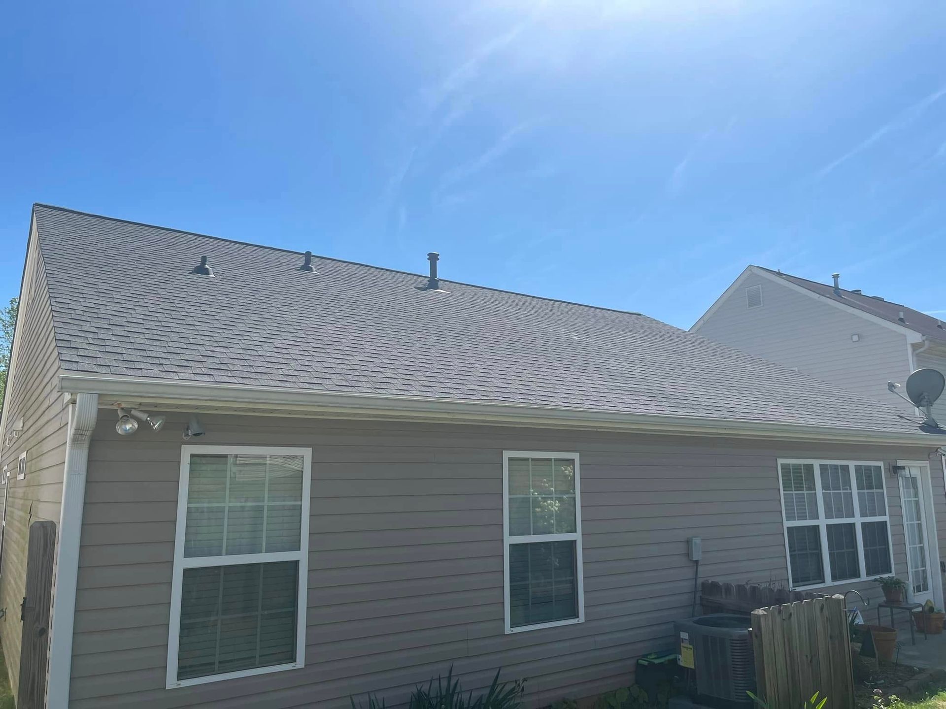 Side of a beige house with white-trimmed windows and a shingled roof under a bright blue sky