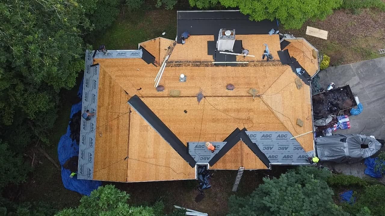 Aerial view of a house roof under construction with plywood decking and scattered tools, surrounded by trees.