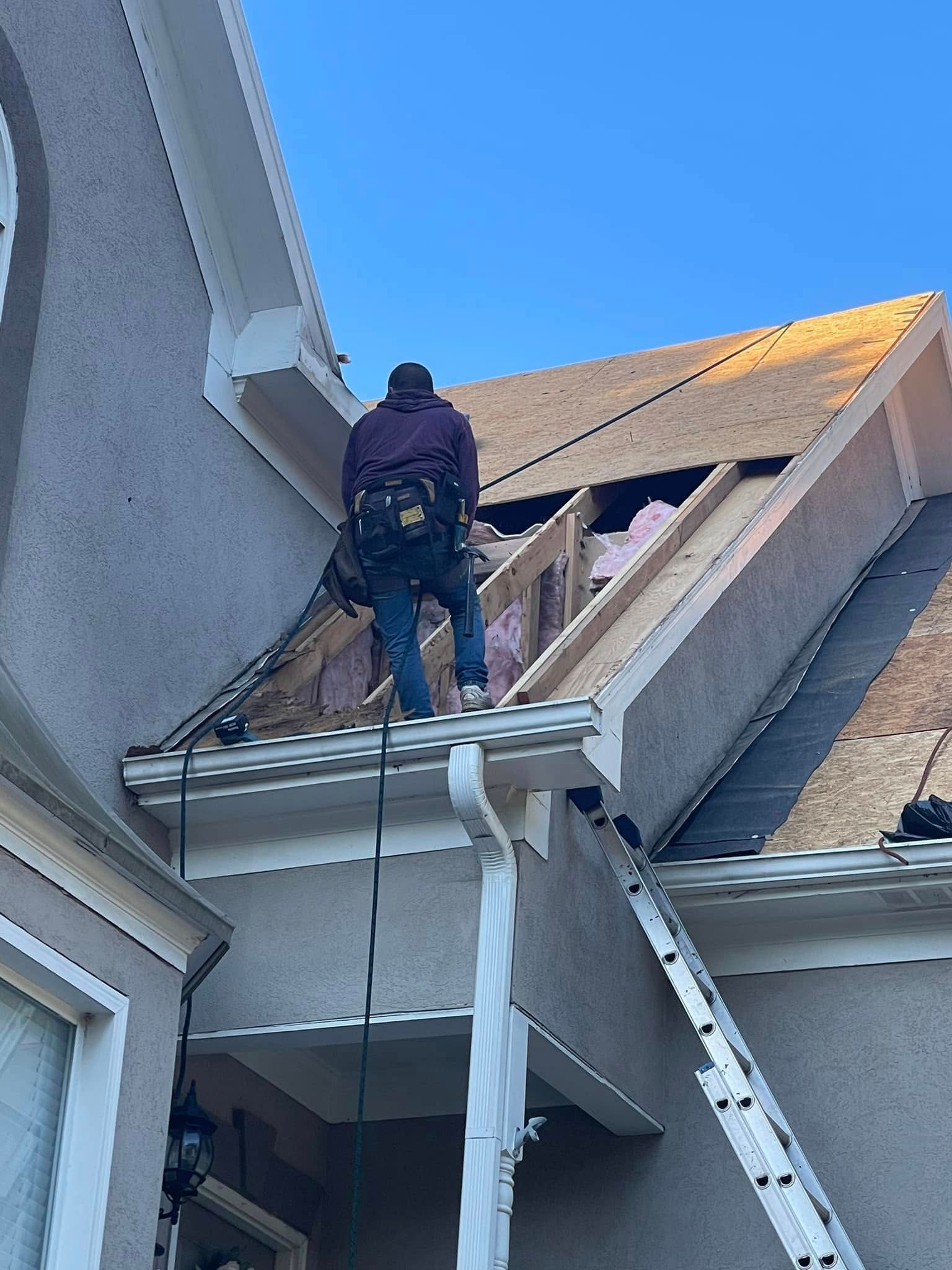 Worker on a roof with ladder, repairing damage near a house wall and eaves.