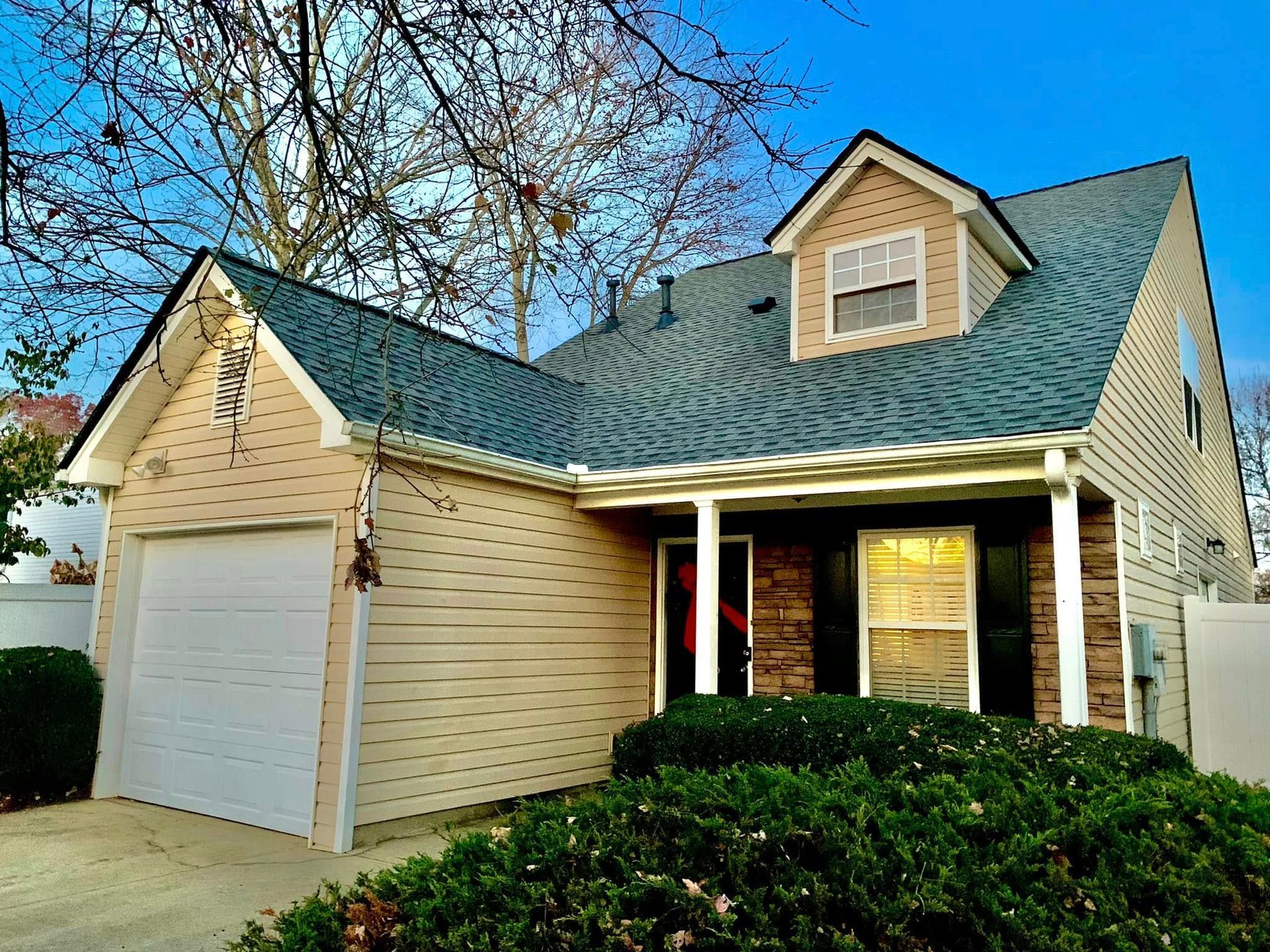 Suburban beige house with blue roof, white garage, and front porch behind green shrubs