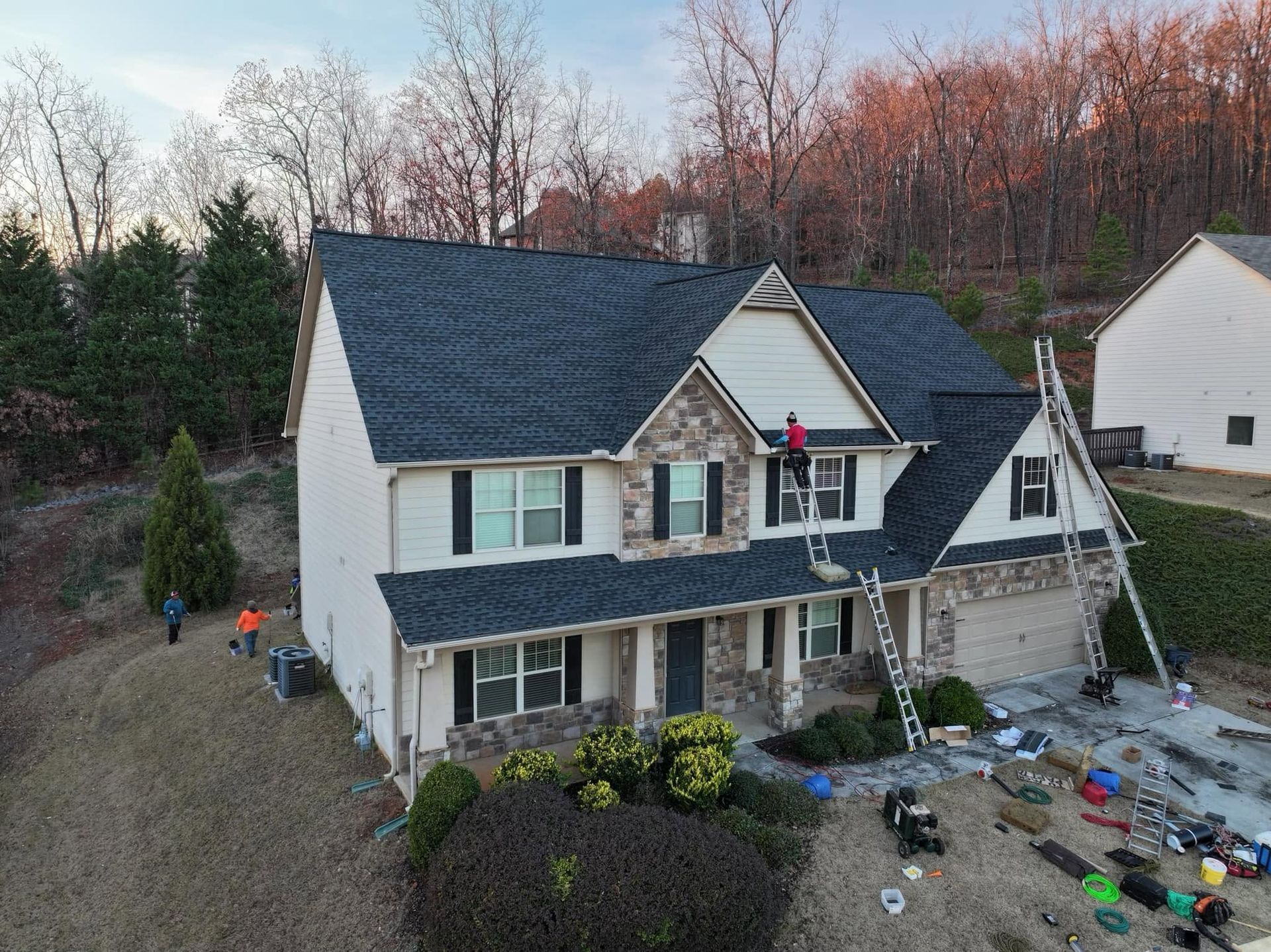 Two-story gray house with black roof and ladder, surrounded by yard tools and fall trees at dusk