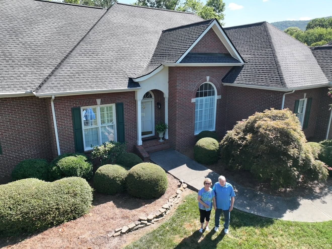 Two people standing on a walkway in front of a brick house with trimmed bushes.