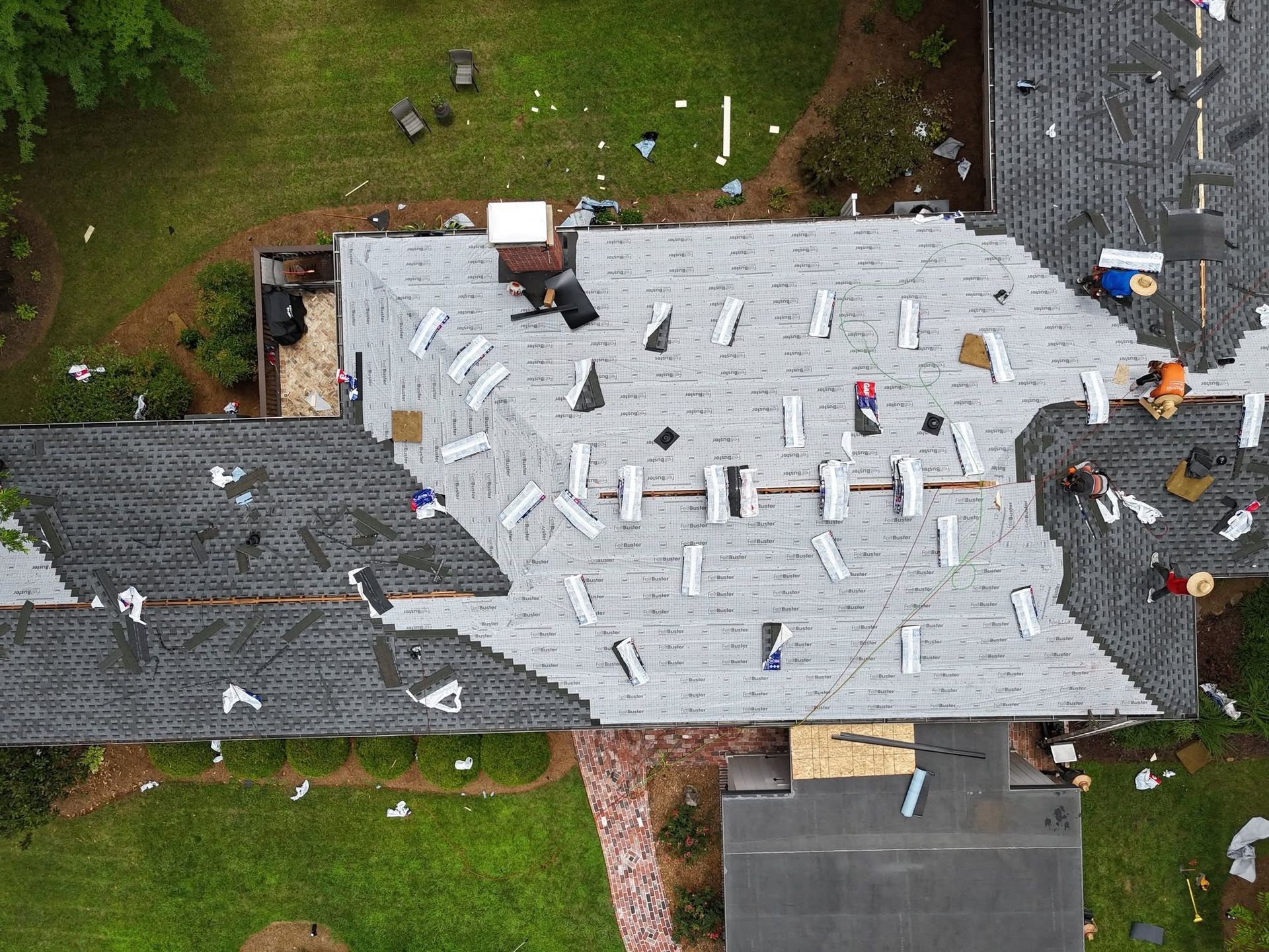 Aerial view of a house roof with gray shingles, white underlayment, and scattered roofing materials during repairs