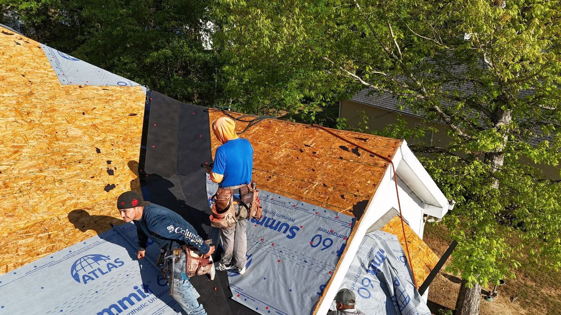Two workers installing roofing felt on a steep roof, with trees in the background.