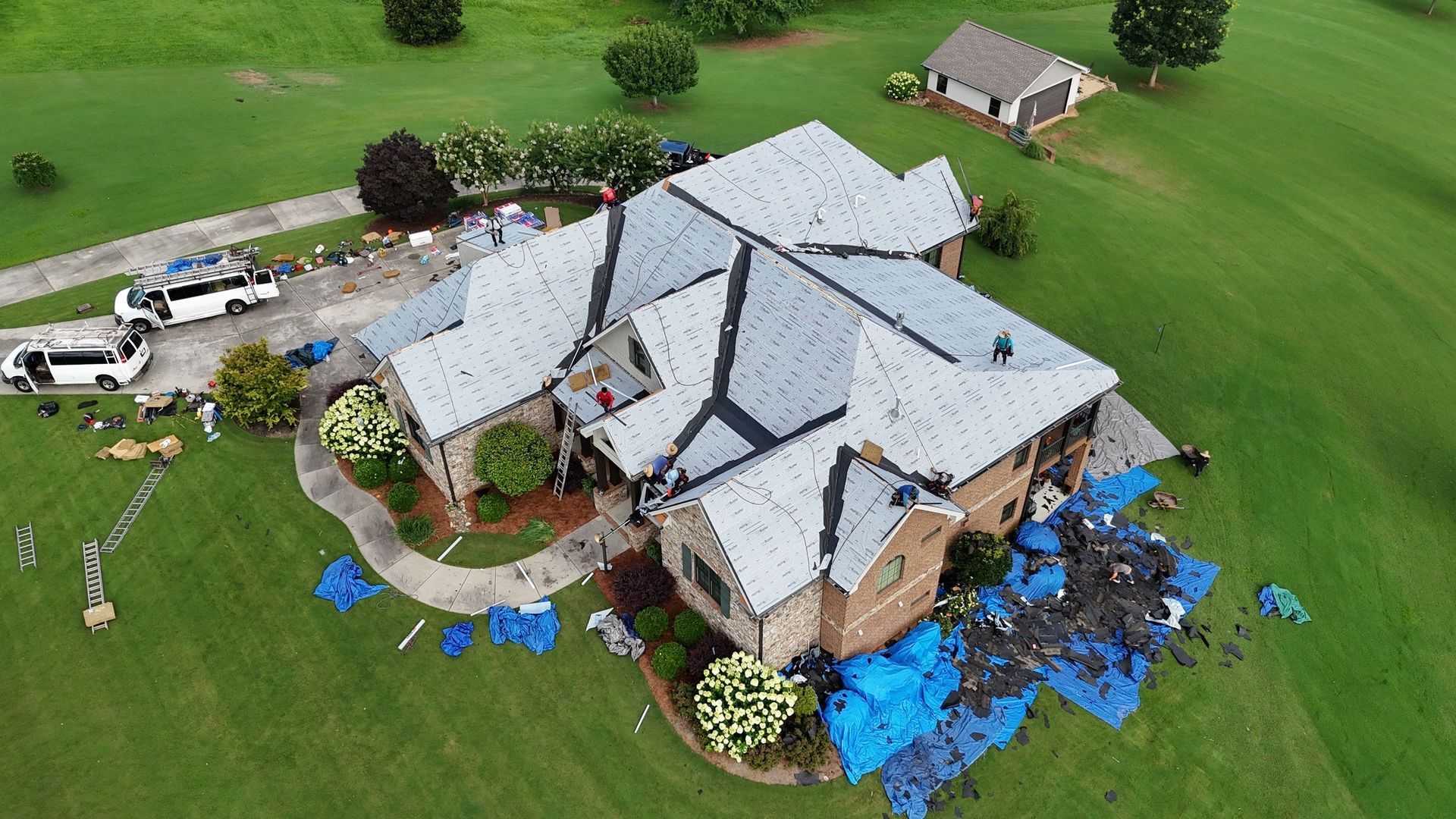 Aerial view of a damaged house with tarp-covered roof and debris scattered around a green yard.