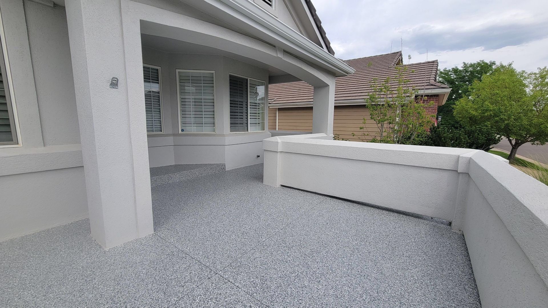 White house porch with covered patio, gravel ground, and low wall beside a tree-lined driveway
