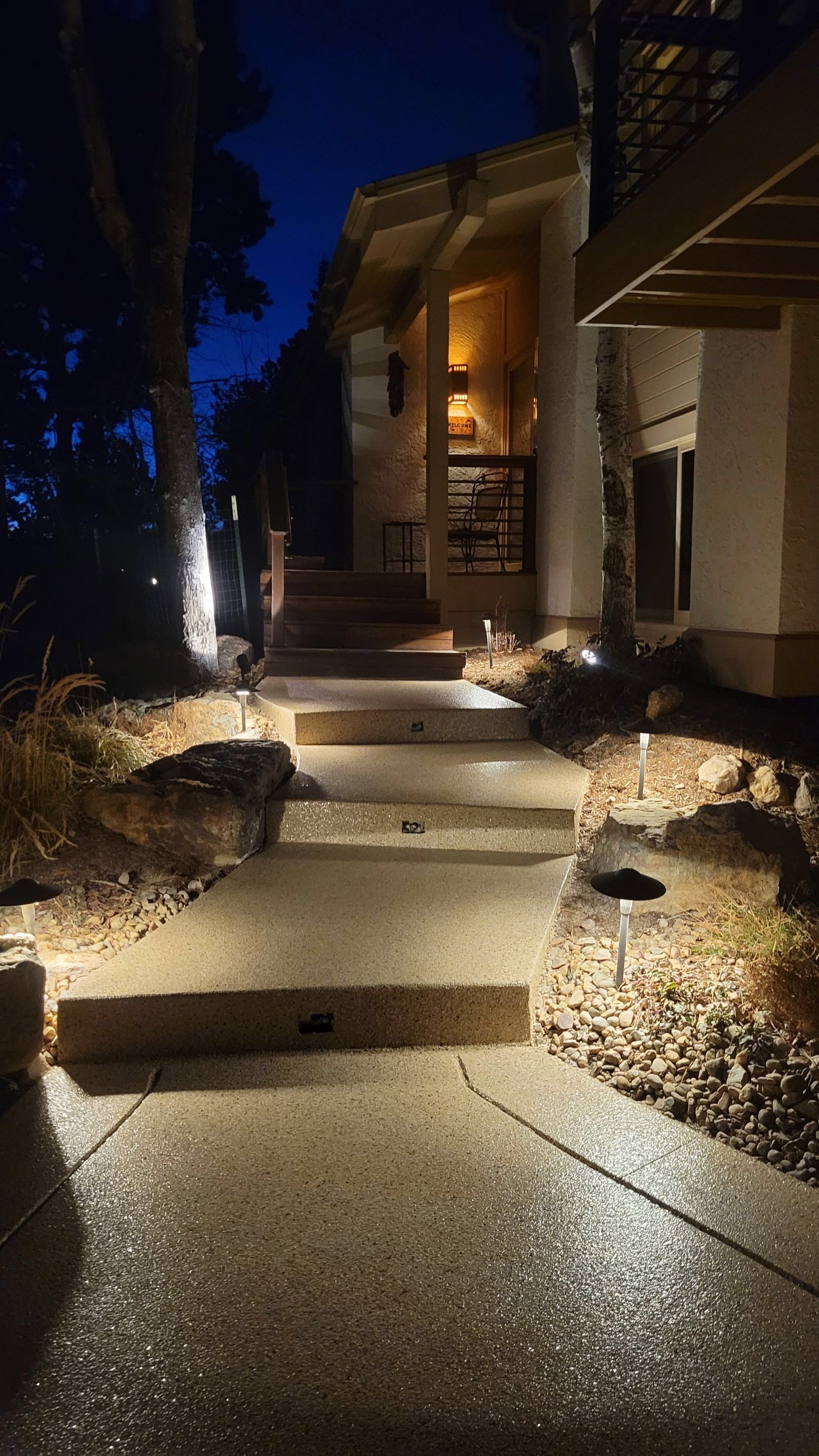 Night-lit stone walkway beside a house, lined with shrubs and warm garden lights.