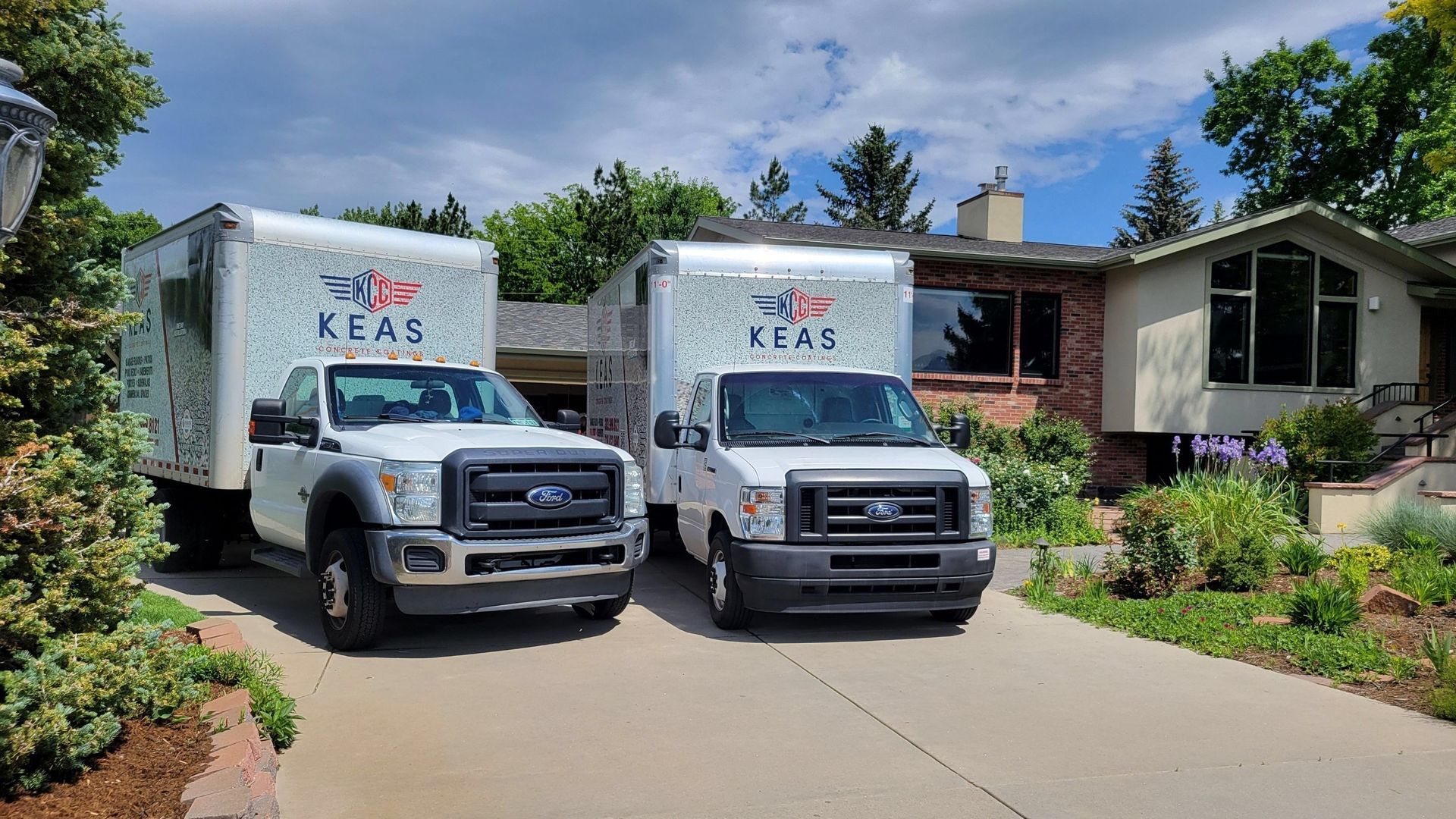Two white KES box trucks parked in a driveway beside a suburban house and garden.