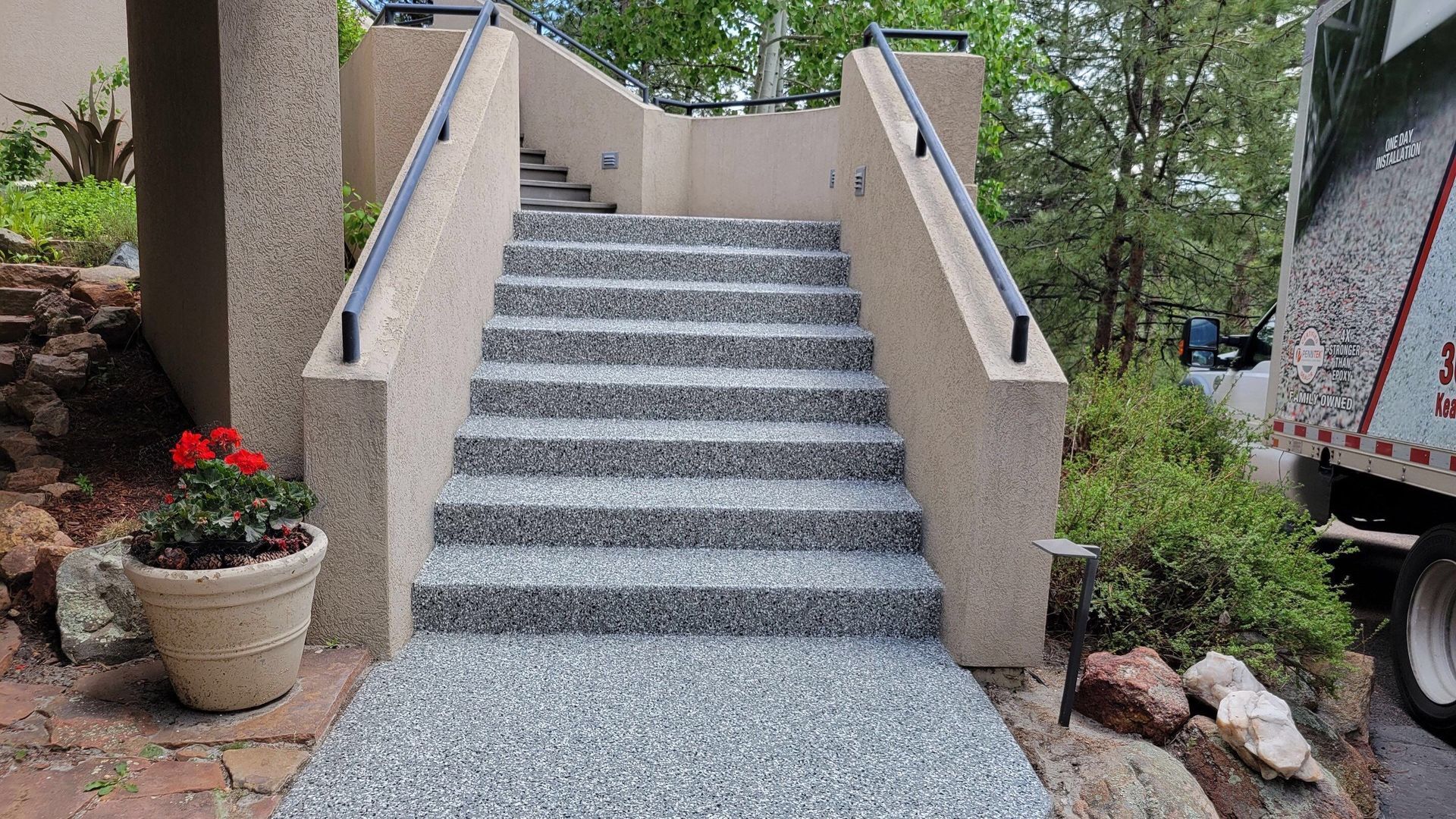 Stone staircase with beige walls and gray steps leading up outdoors, flanked by potted flowers and trees