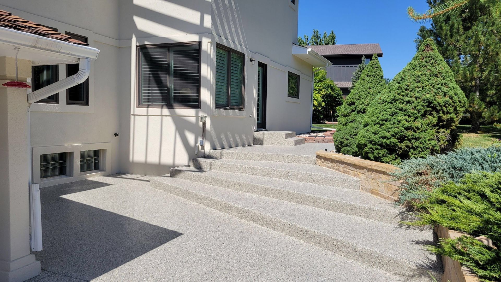 Sunlit modern house patio with concrete steps, windows, and landscaped shrubs