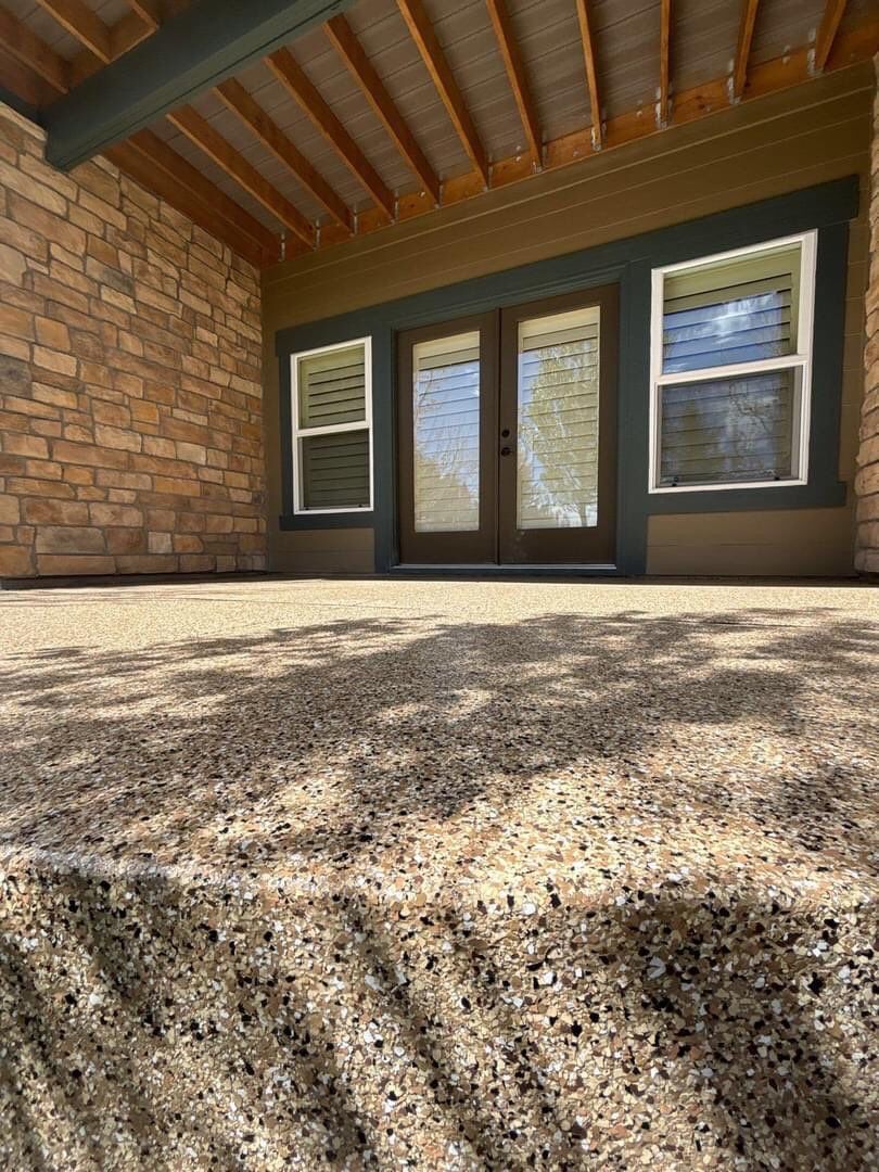 Covered patio with stone wall, wood ceiling, double glass doors, and two windows in bright sunlight.