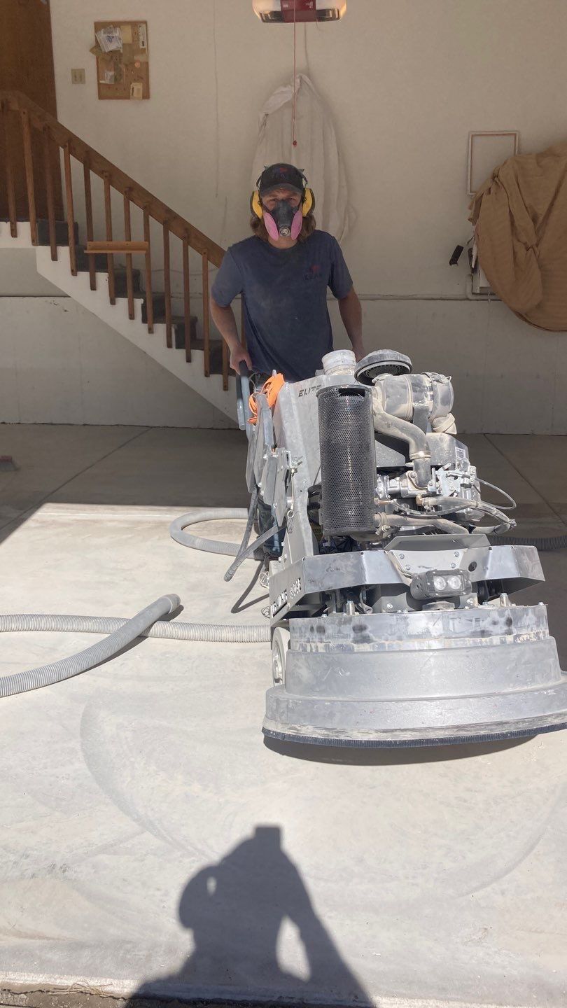 Worker operating a concrete grinder in a garage, with dust on the floor and stairs in the background