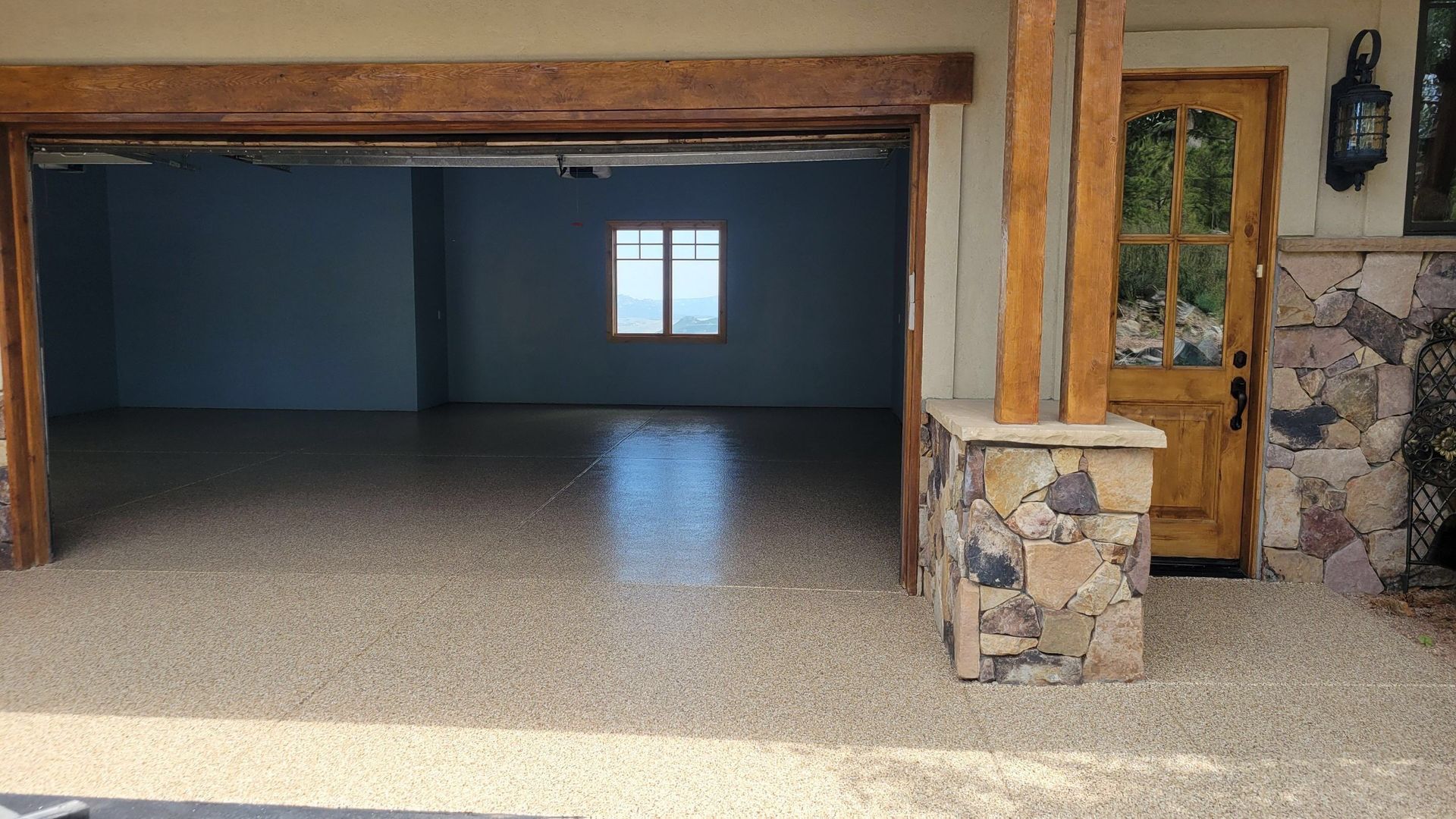 Open garage with blue interior, speckled floor, wood beams, small window, and stone pillar with wooden door.