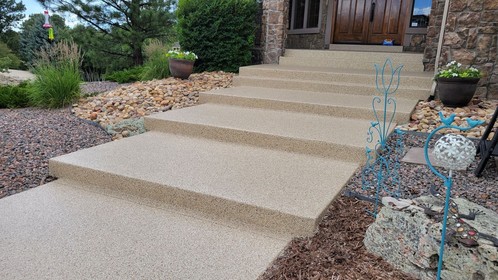 Stone front walkway with wide steps leading to a wooden front door, bordered by garden beds and potted plants