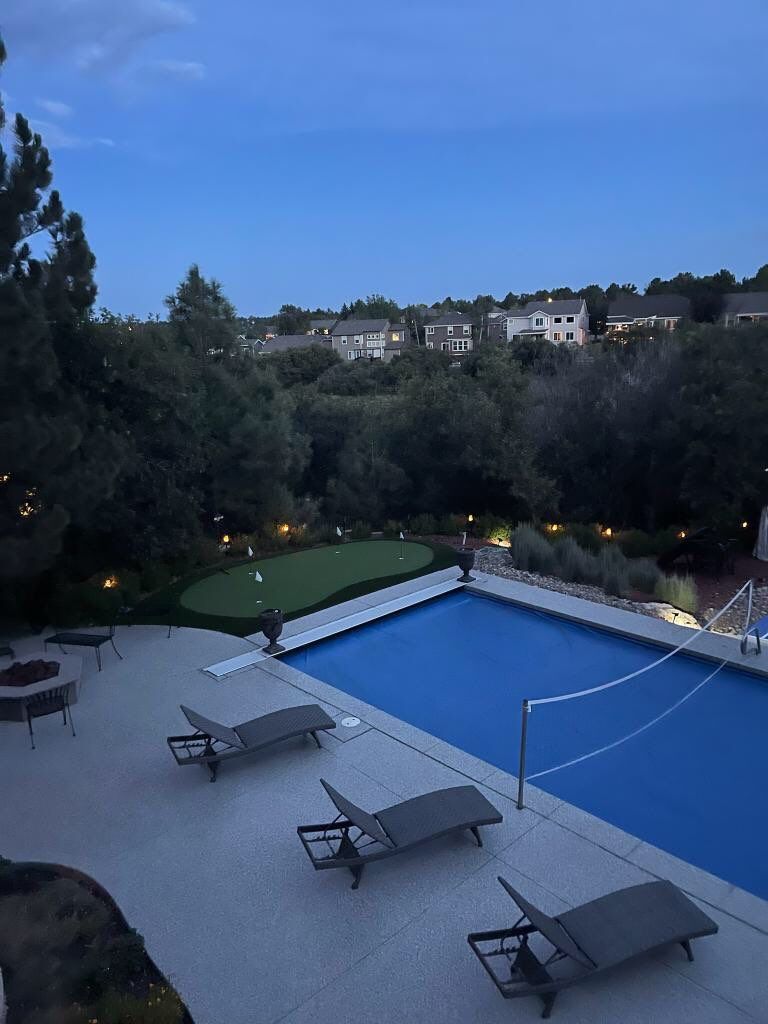 Outdoor pool at dusk with blue court, lounge chairs, and hillside homes beyond trees