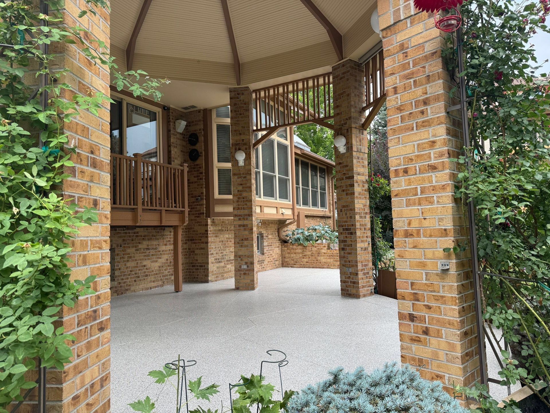 Covered brick patio with wooden porch, arched columns, and gravel courtyard surrounded by greenery