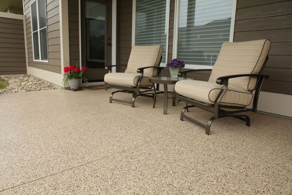 Two beige lounge chairs on a pebble patio beside a house, with potted flowers and sliding glass doors.