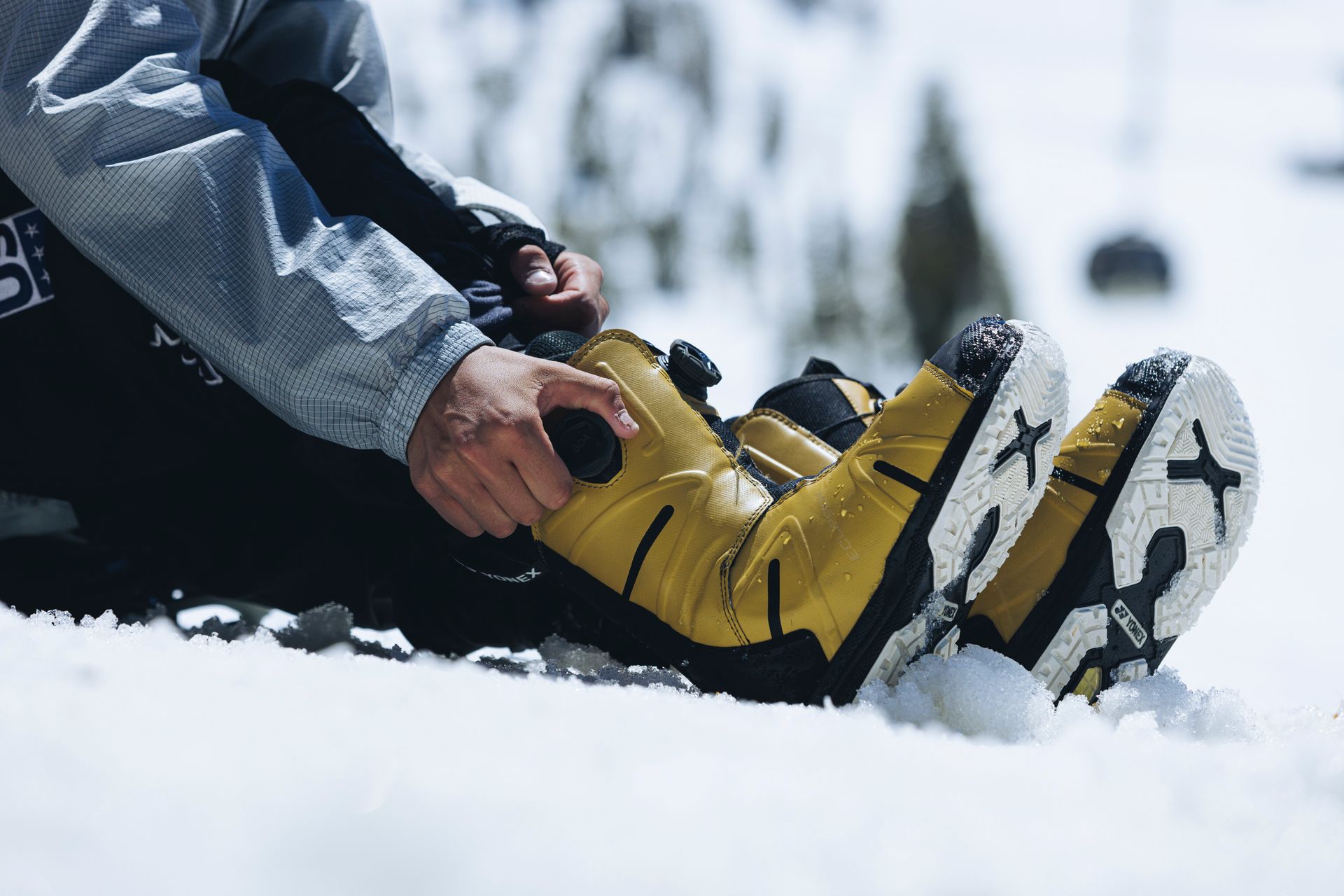 Eine Person sitzt im Schnee und rückt ihre gelben und schwarzen Schneestiefel zurecht.