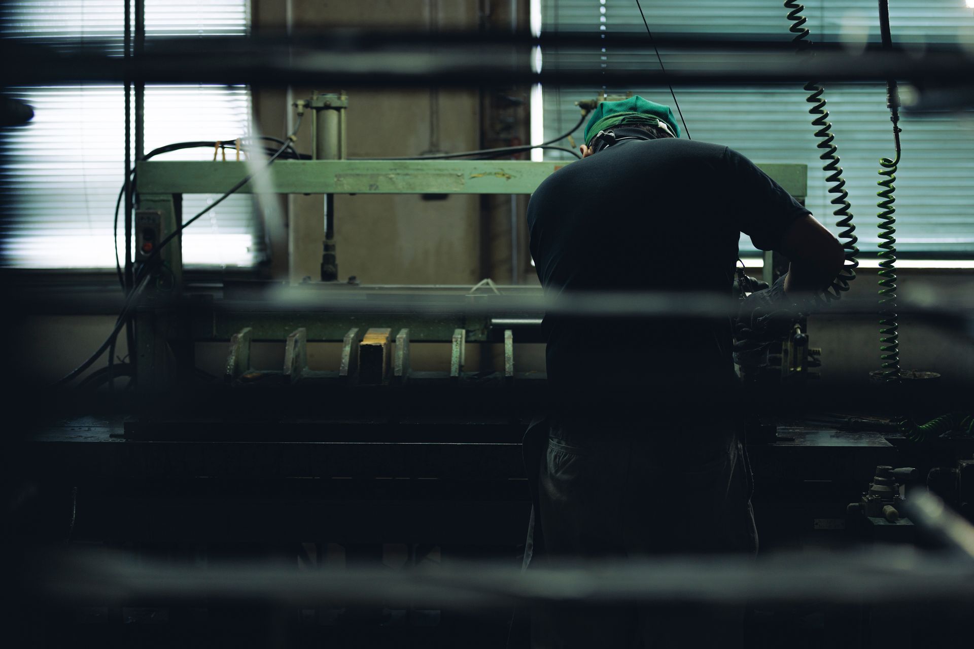 Arbeiter bedient Maschinen in einer düsteren Industrieumgebung. Dunkle Kleidung, grüne Kopfbedeckung, arbeitet an einer langen Maschine.
