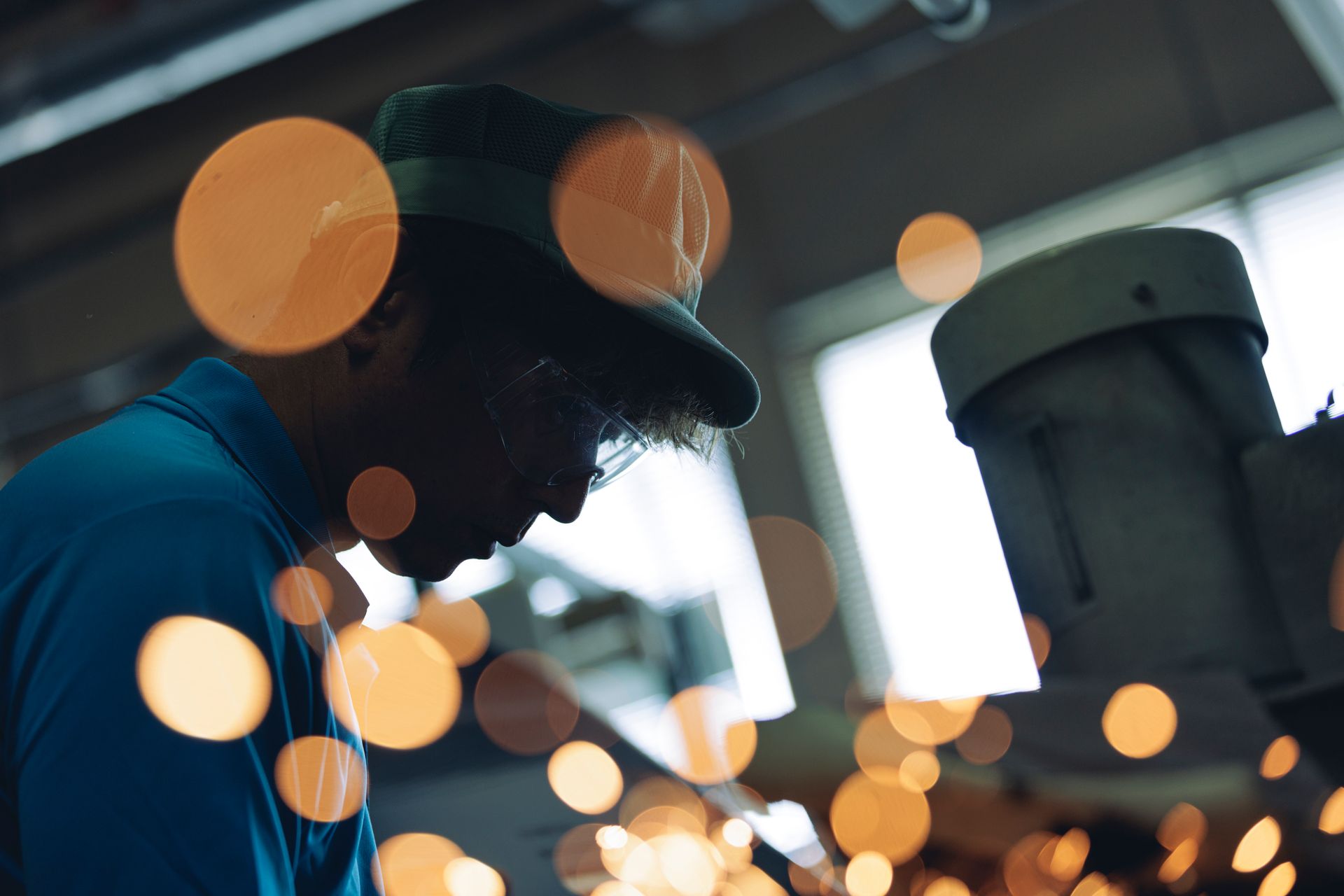 Eine Person mit Hut arbeitet in einer Werkstatt an Maschinen. Orangefarbene Funken und Bokeh-Licht.