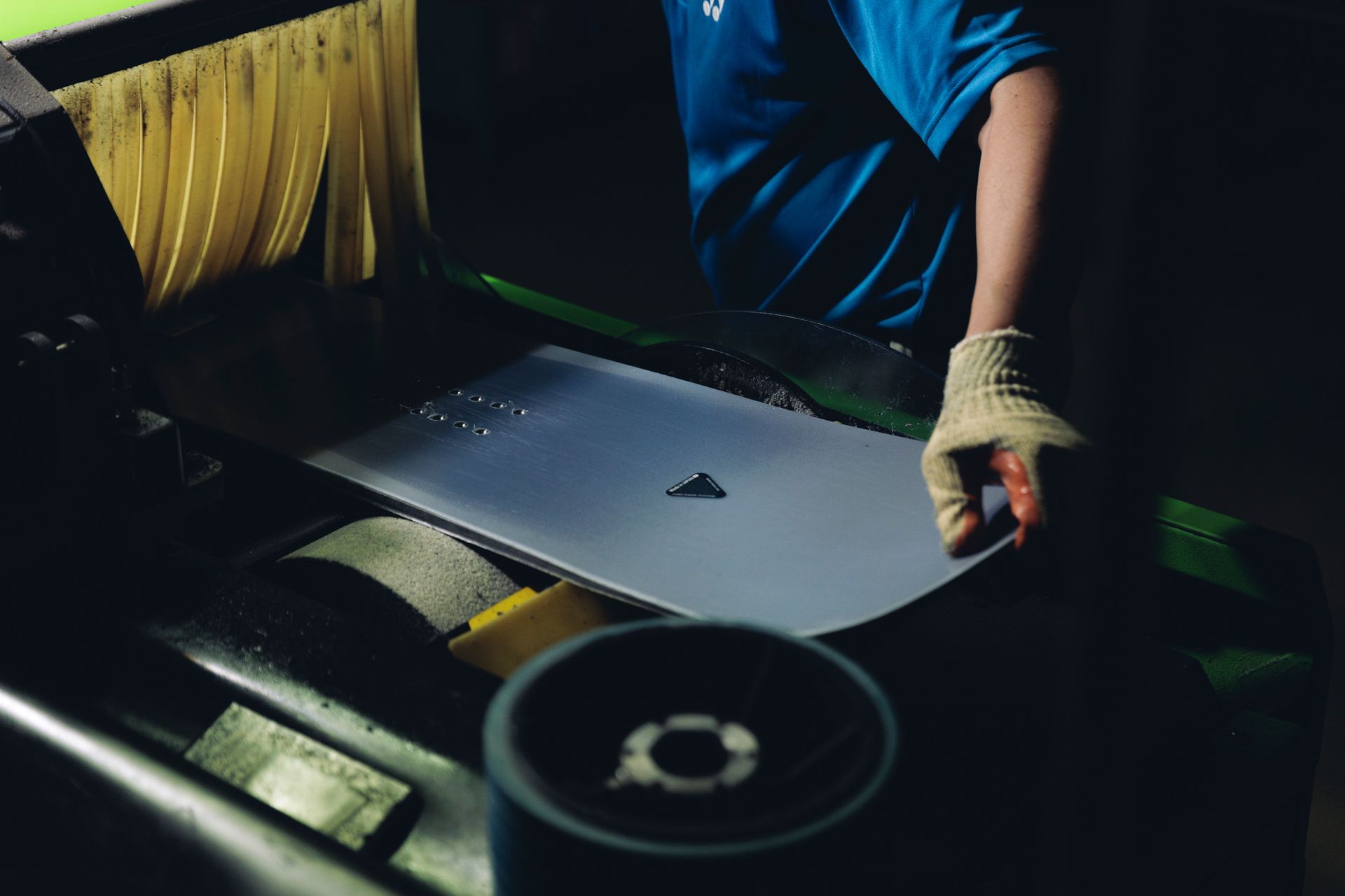 Ein Arbeiter in blauem Hemd und Handschuh hantiert mit einem Metallblech an einer dunklen Industriemaschine.