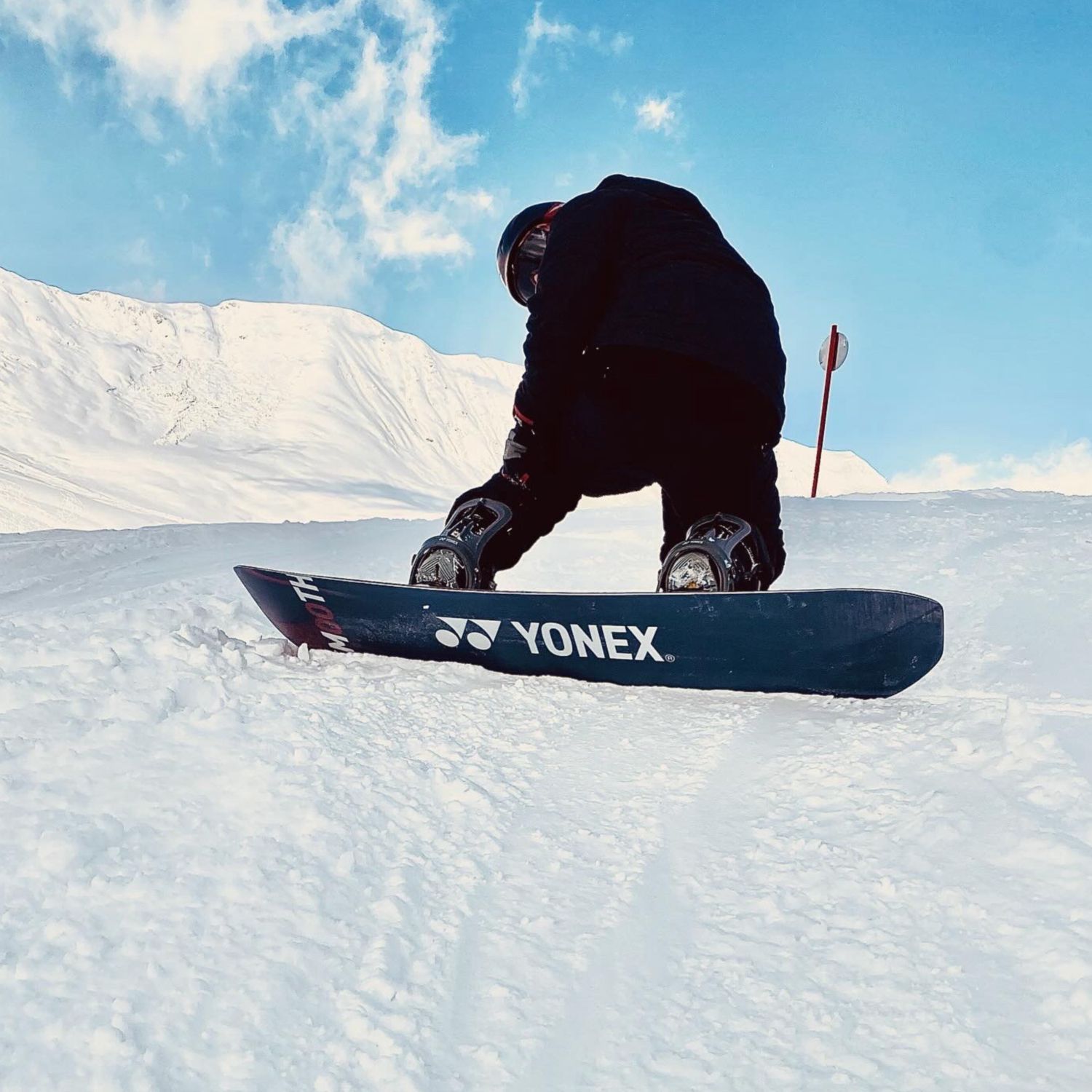 Ein Snowboarder hockte auf einem Snowboard mit YONEX-Logo an einem schneebedeckten Berghang unter blauem Himmel.