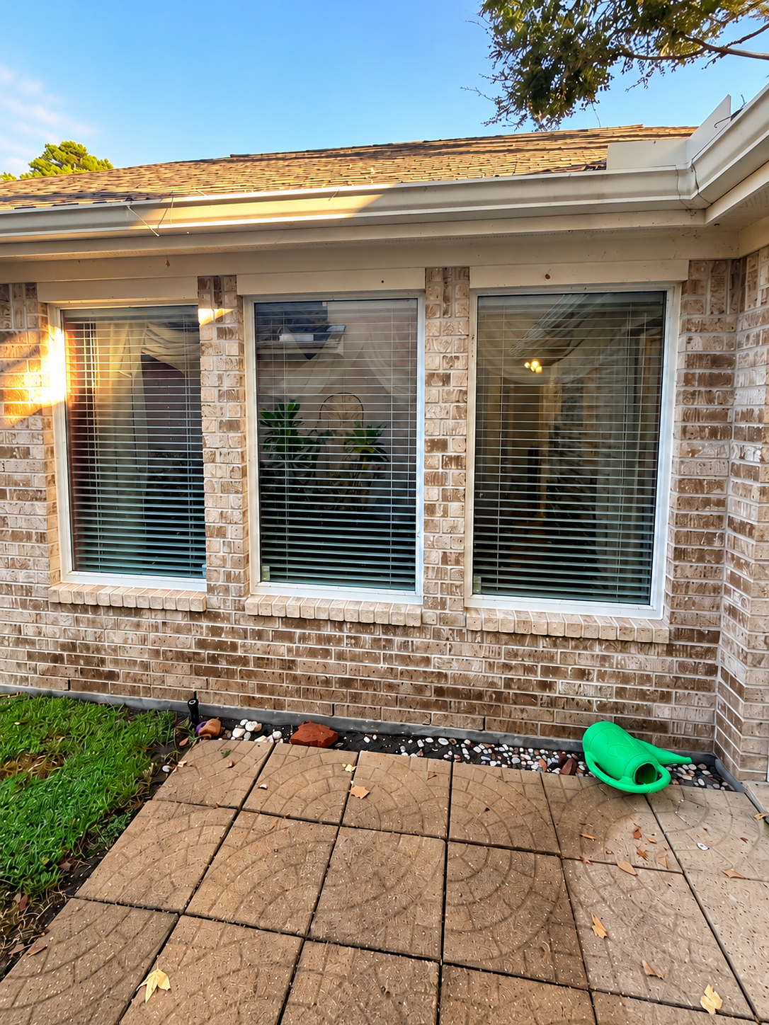 Brick house exterior with three windows, a tiled patio, and a green watering can on the ground