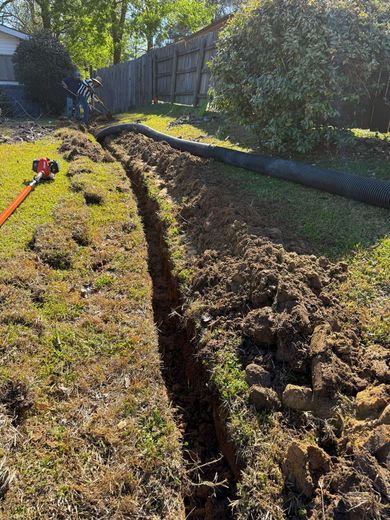 Deep trench dug in a grassy yard beside a fence, with a shovel on the left.