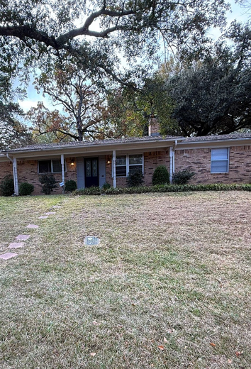 Single-story brick house with a front porch and overgrown yard under large trees.