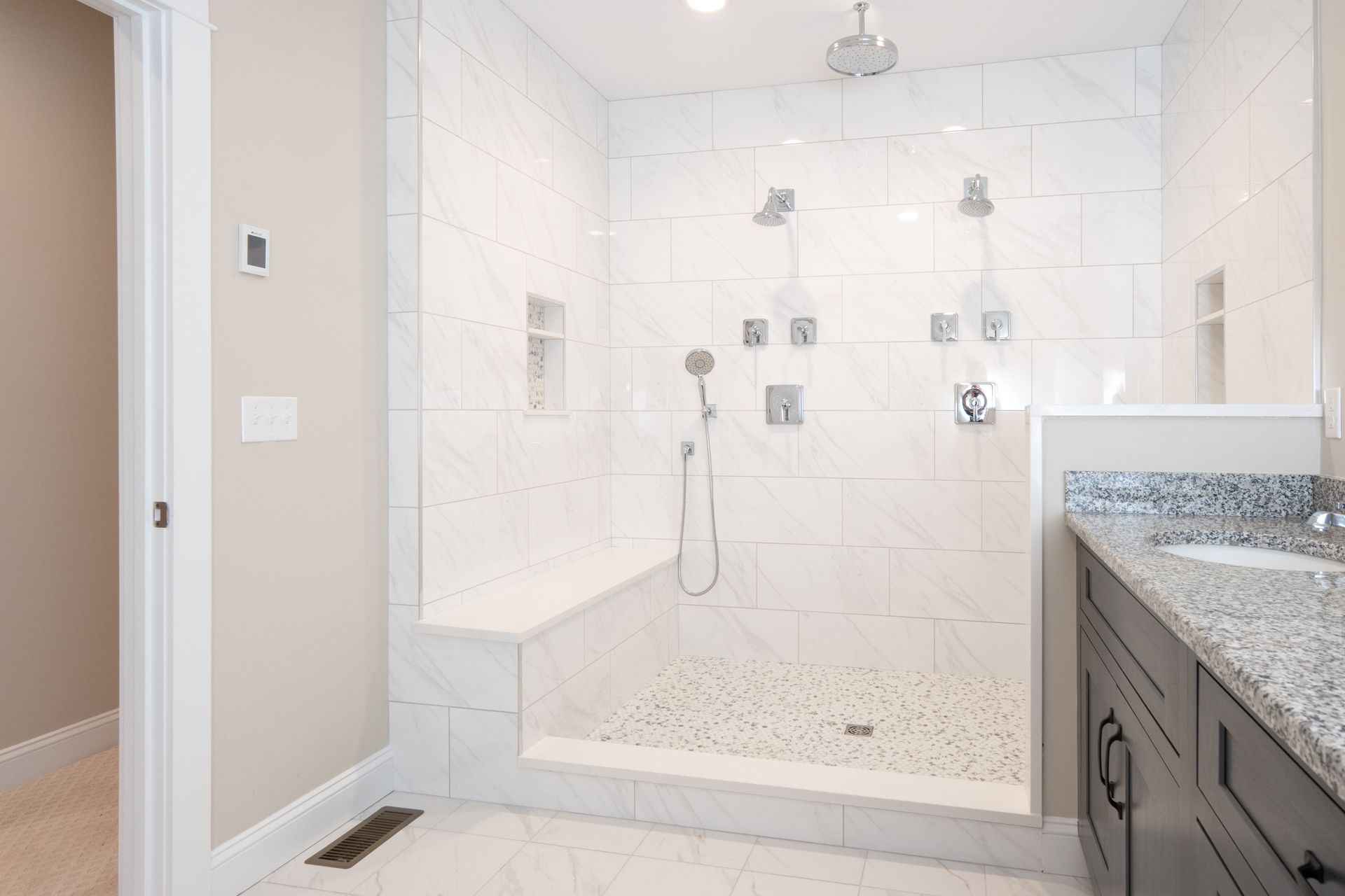 A walk-in shower with white tiled walls, a built-in bench, dual showerheads, and a stone-tiled floor next to a vanity.