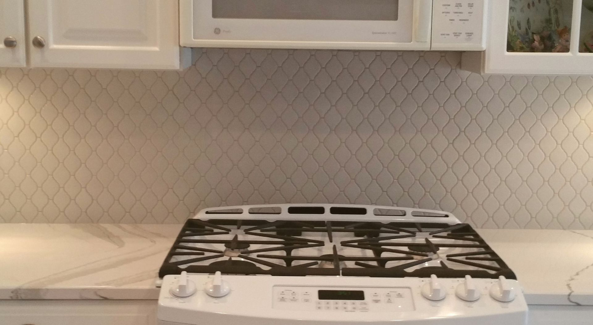 A white kitchen gas stove and marble countertop with a white patterned tile backsplash beneath a microwave.
