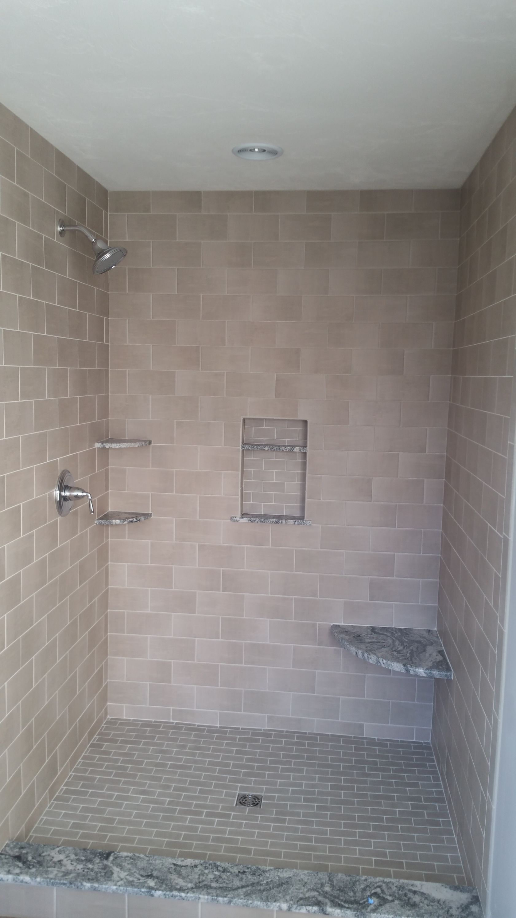 A walk-in shower with tan subway tile walls, a built-in recessed shelf, a stone corner seat, and a gray pebble tile floor.
