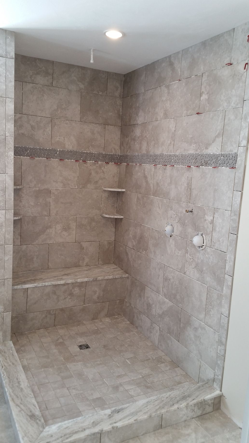 A neutral-toned tiled walk-in shower featuring a built-in stone bench, two corner shelves, and a floor-level drain.