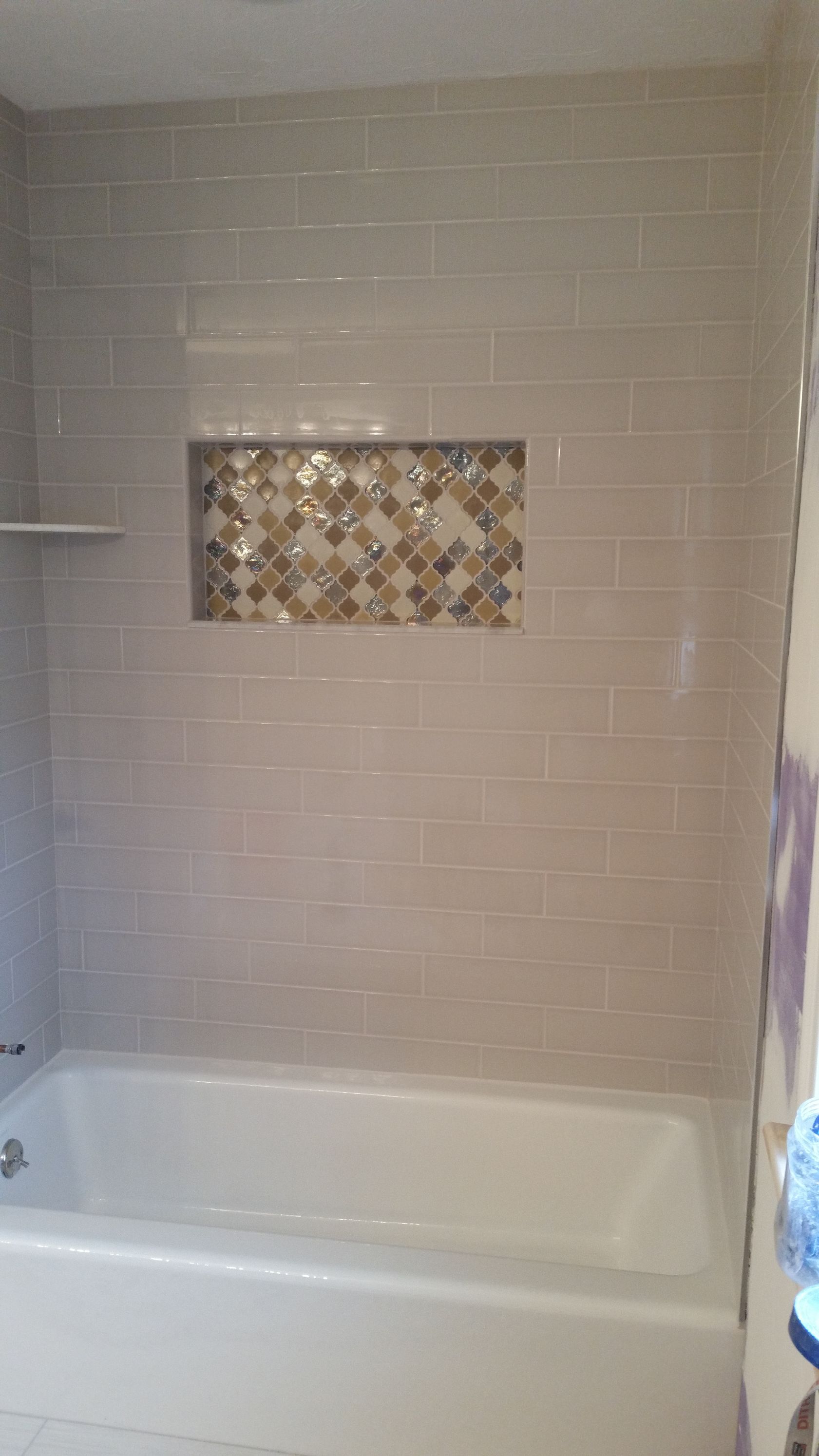 A white bathtub with a beige tiled shower wall featuring a decorative mosaic-tiled rectangular recessed shelf.