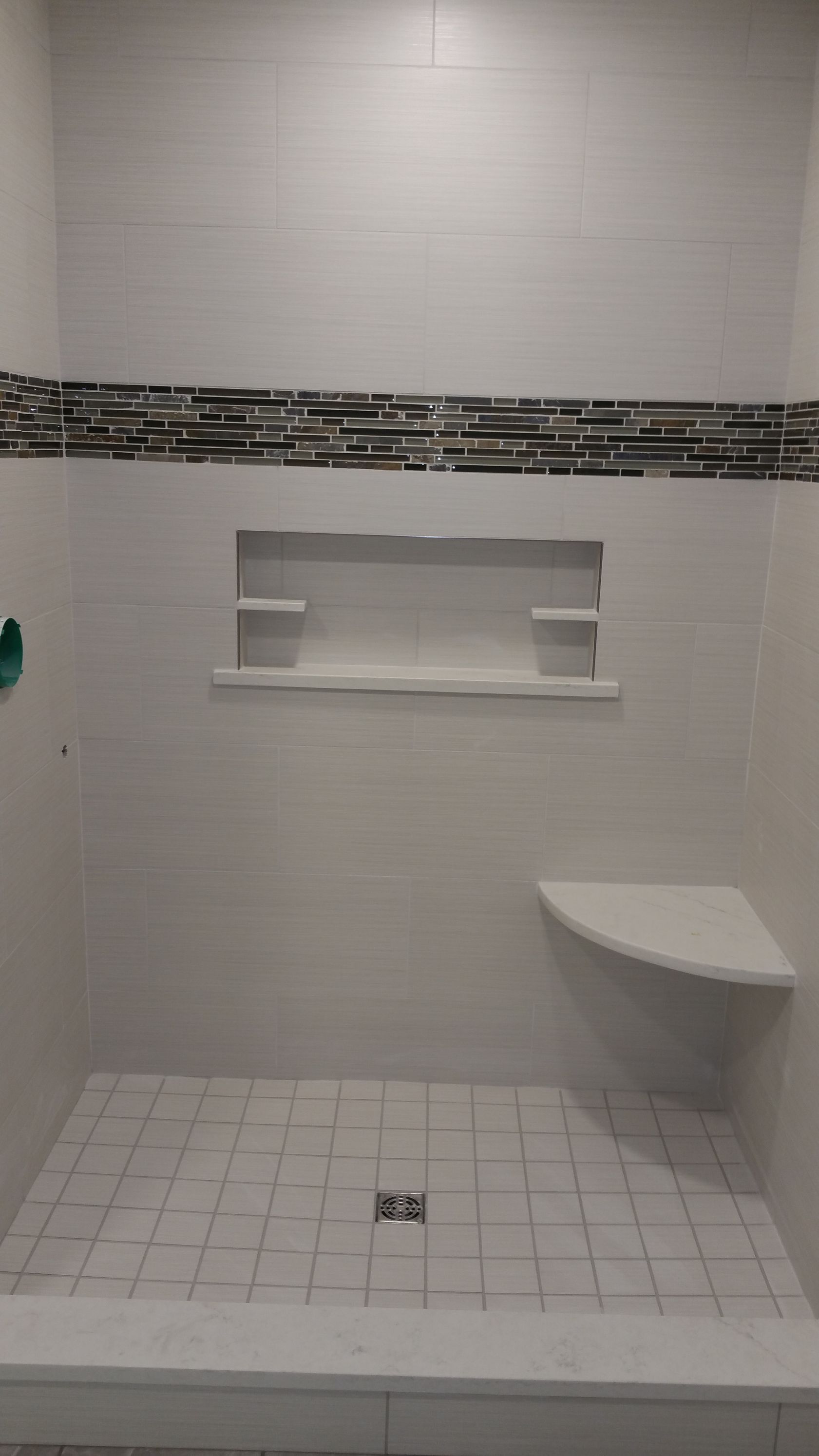 A walk-in shower with white tiled walls and floor, a patterned accent tile stripe, a built-in shelf, and a corner seat.