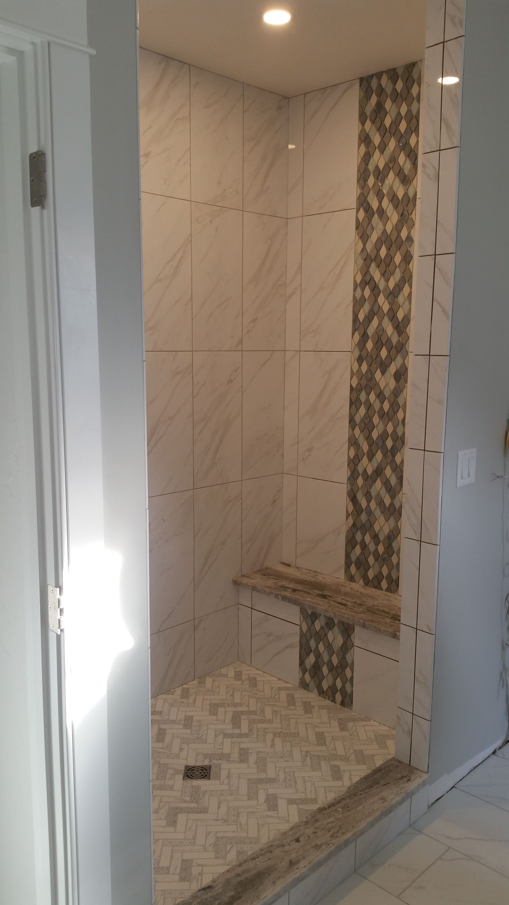 A walk-in shower with light marble-patterned wall tiles, a mosaic tile accent strip, a stone bench, and mosaic floor tile.