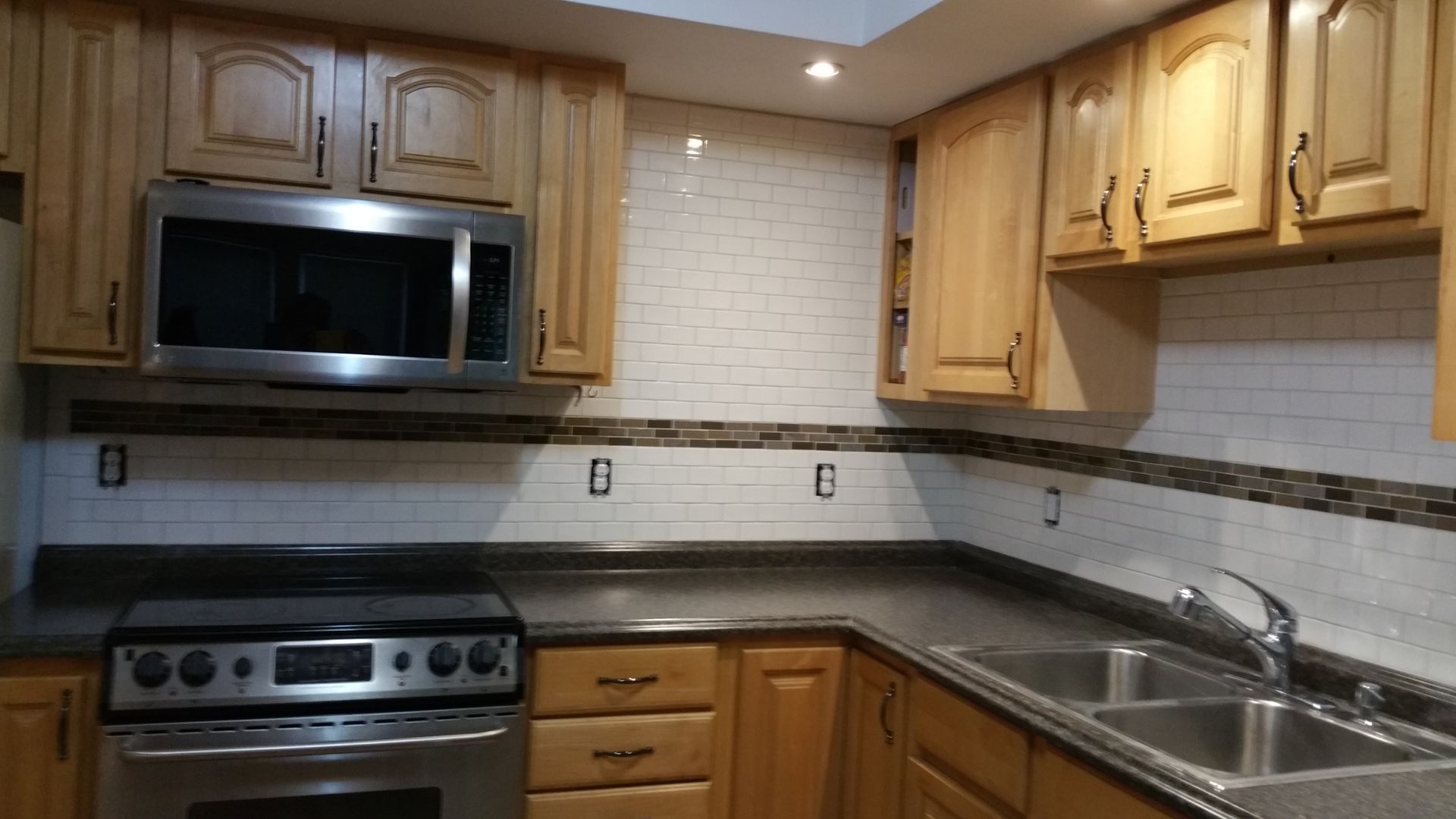 A kitchen with honey-toned cabinets, dark countertops, stainless steel appliances, and a white tiled backsplash.