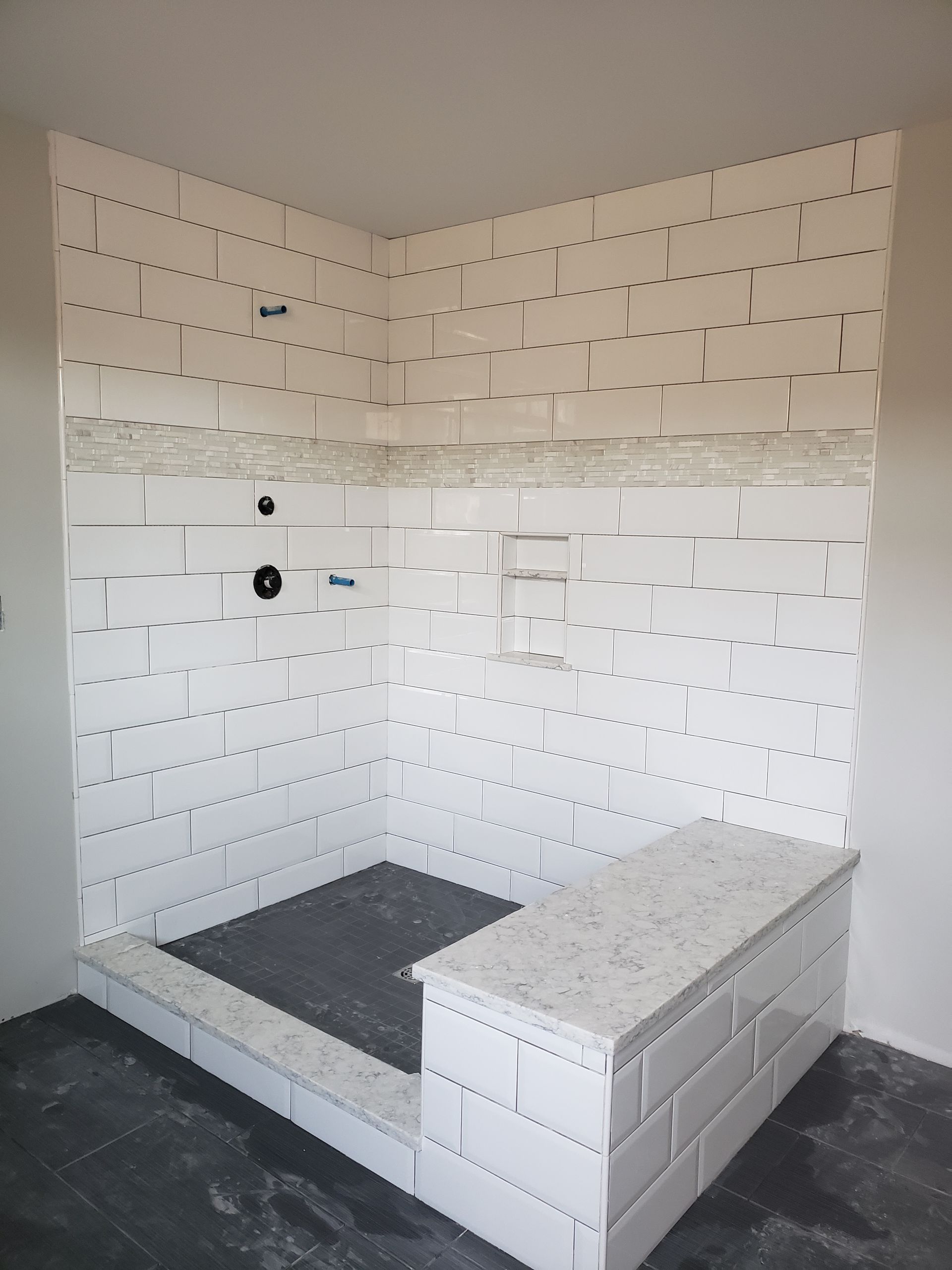 A walk-in shower with white brick-style wall tiles, a light stone bench and accents, and dark grey slate-style floor tiles.