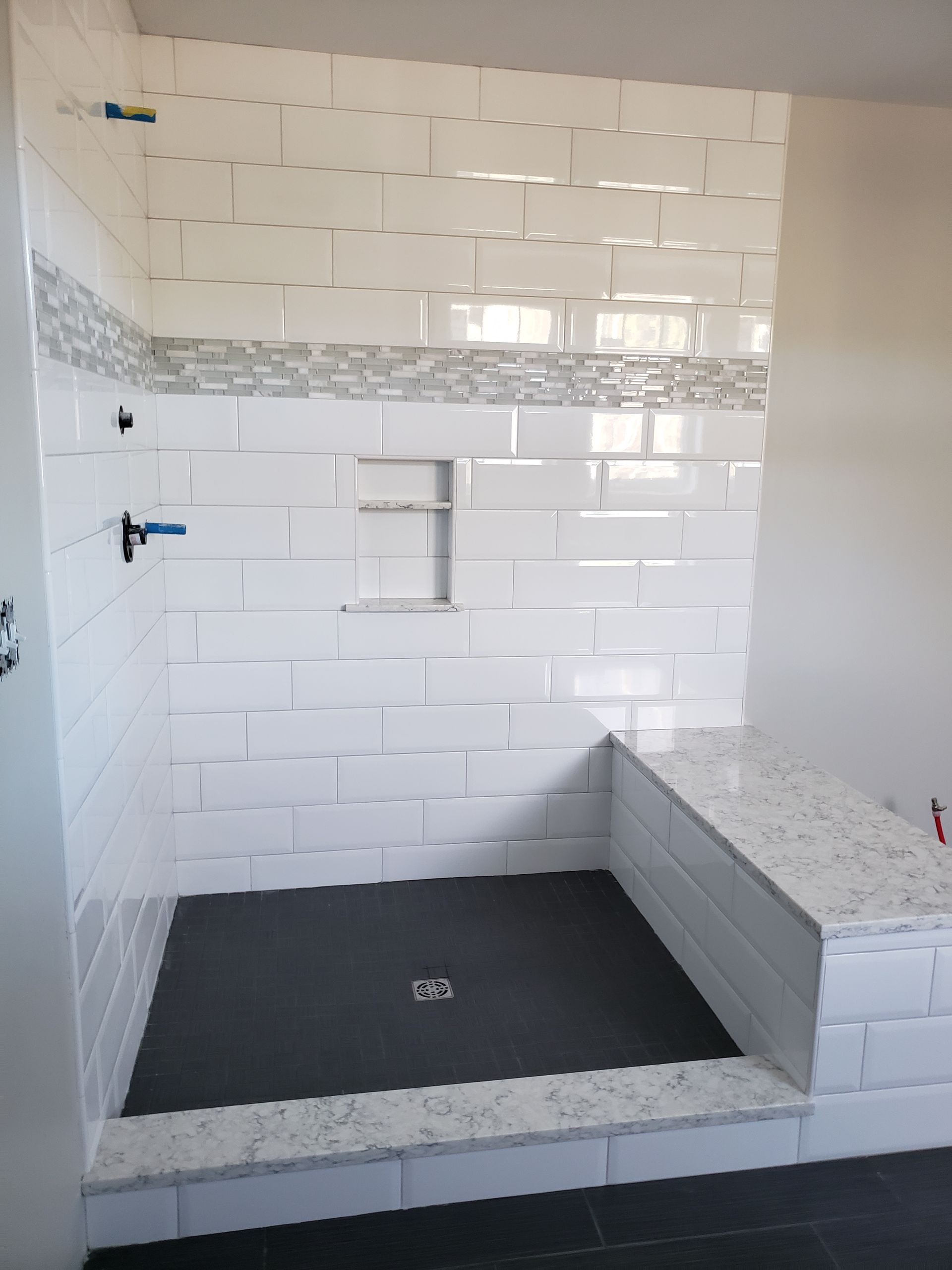 A modern walk-in shower with white subway tiles, a mosaic accent strip, a stone bench, and a dark grey floor.