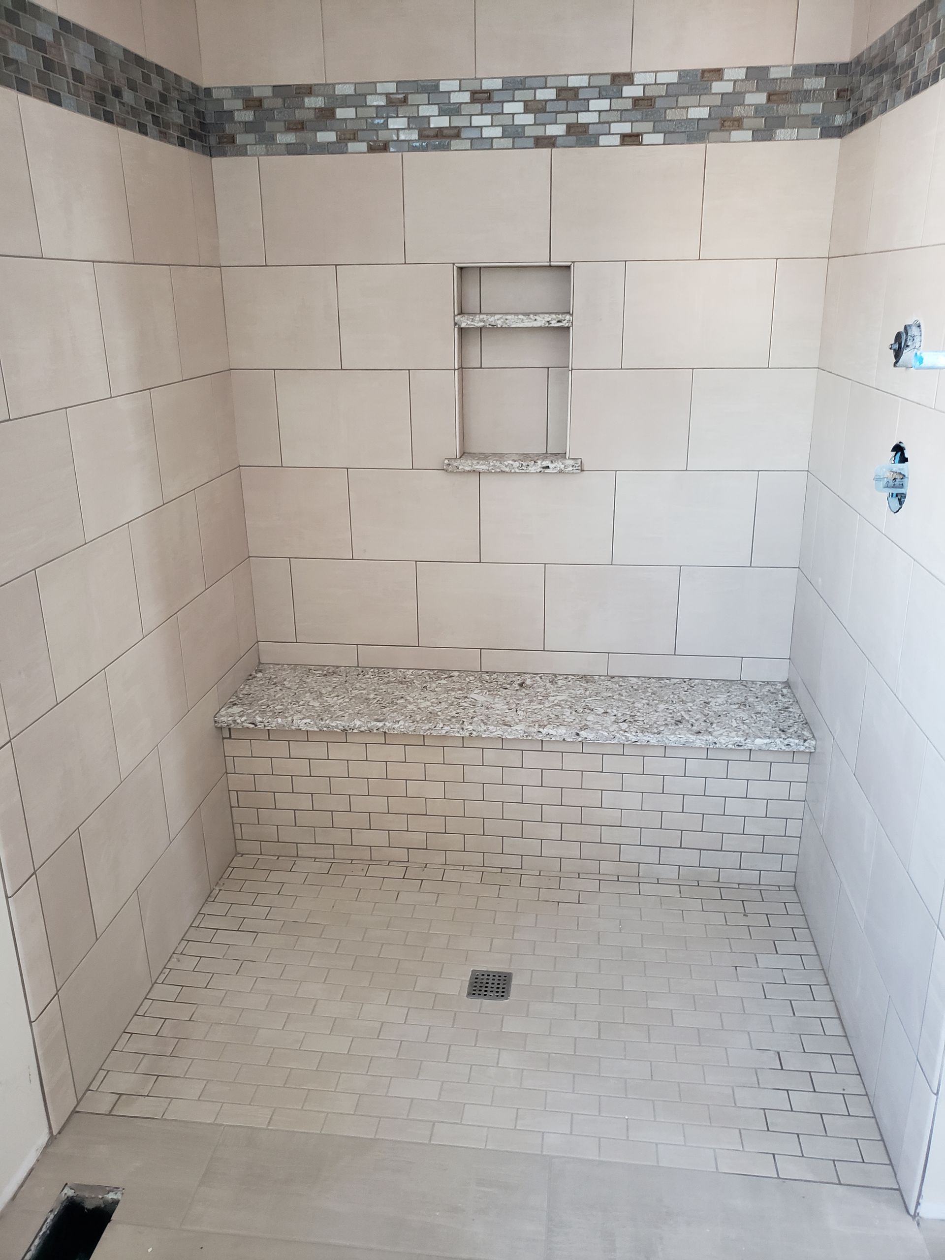 A walk-in shower with beige tiles, a mosaic-tiled accent strip, a built-in bench, and a small recessed wall shelf.