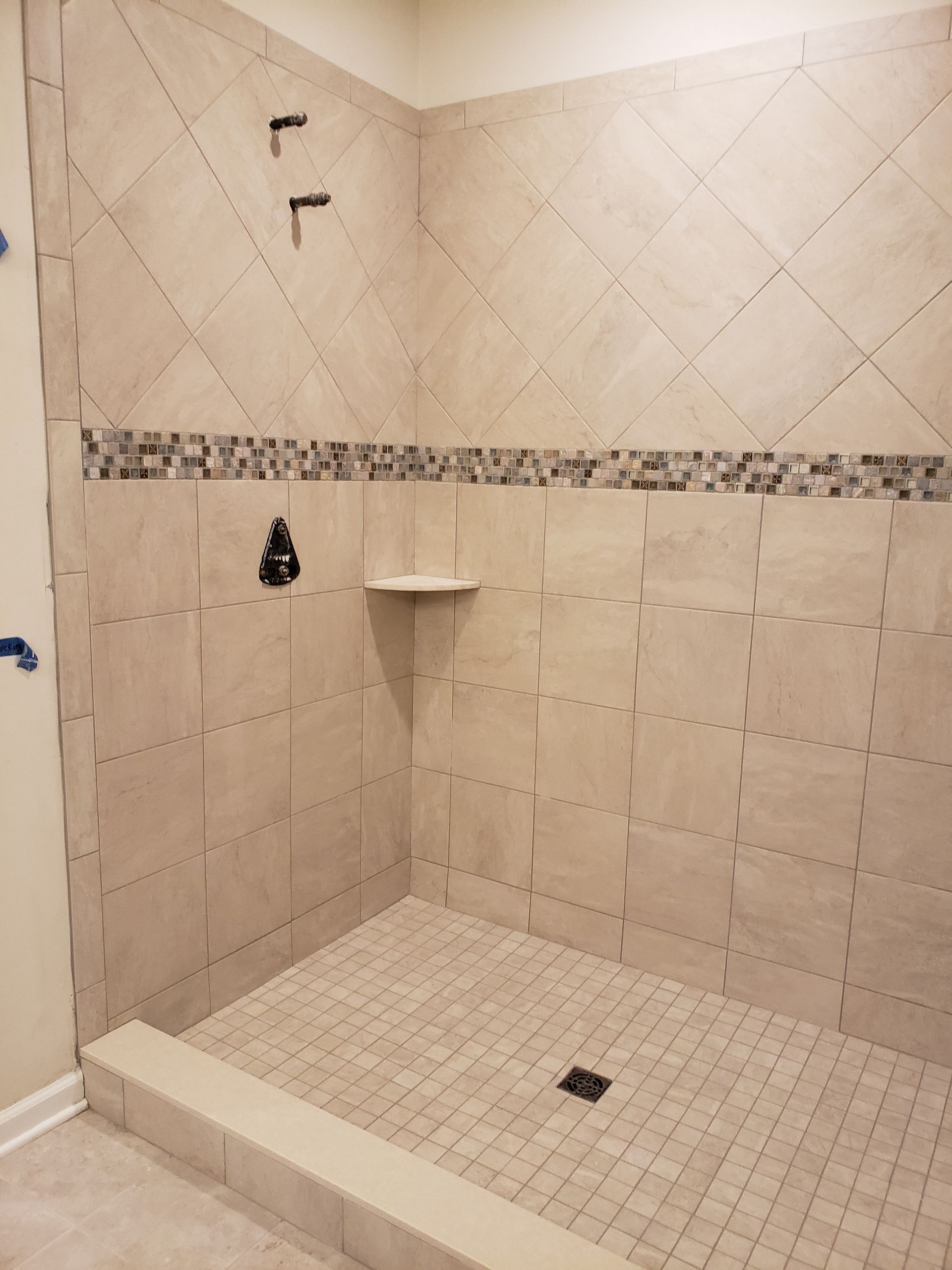 A walk-in shower with beige square tiles, a diagonal tiled upper section, a mosaic accent strip, and a pebble-style floor.