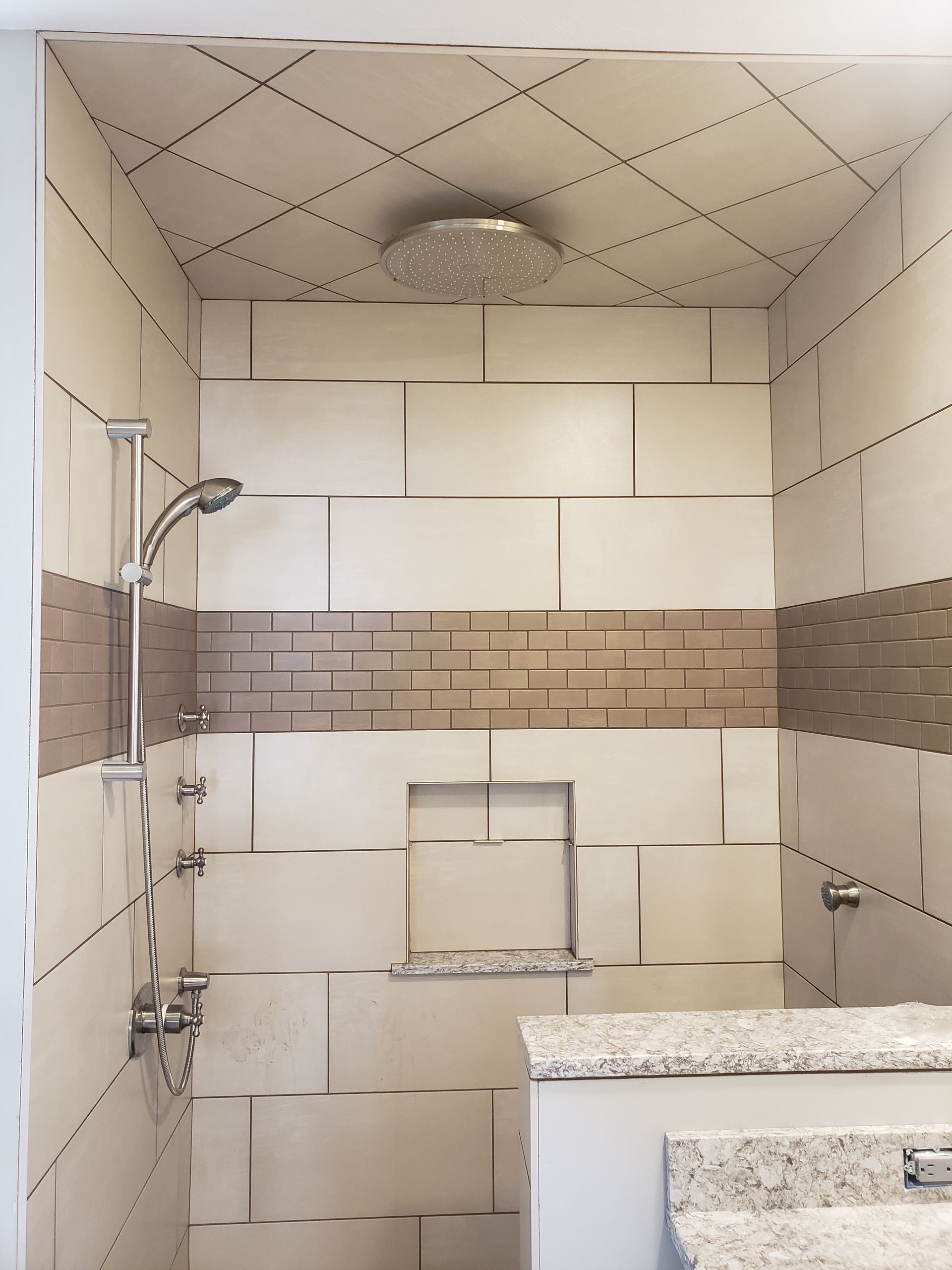 A modern shower with beige tiles, a brown mosaic accent strip, a rainfall showerhead, and a built-in recessed wall shelf.