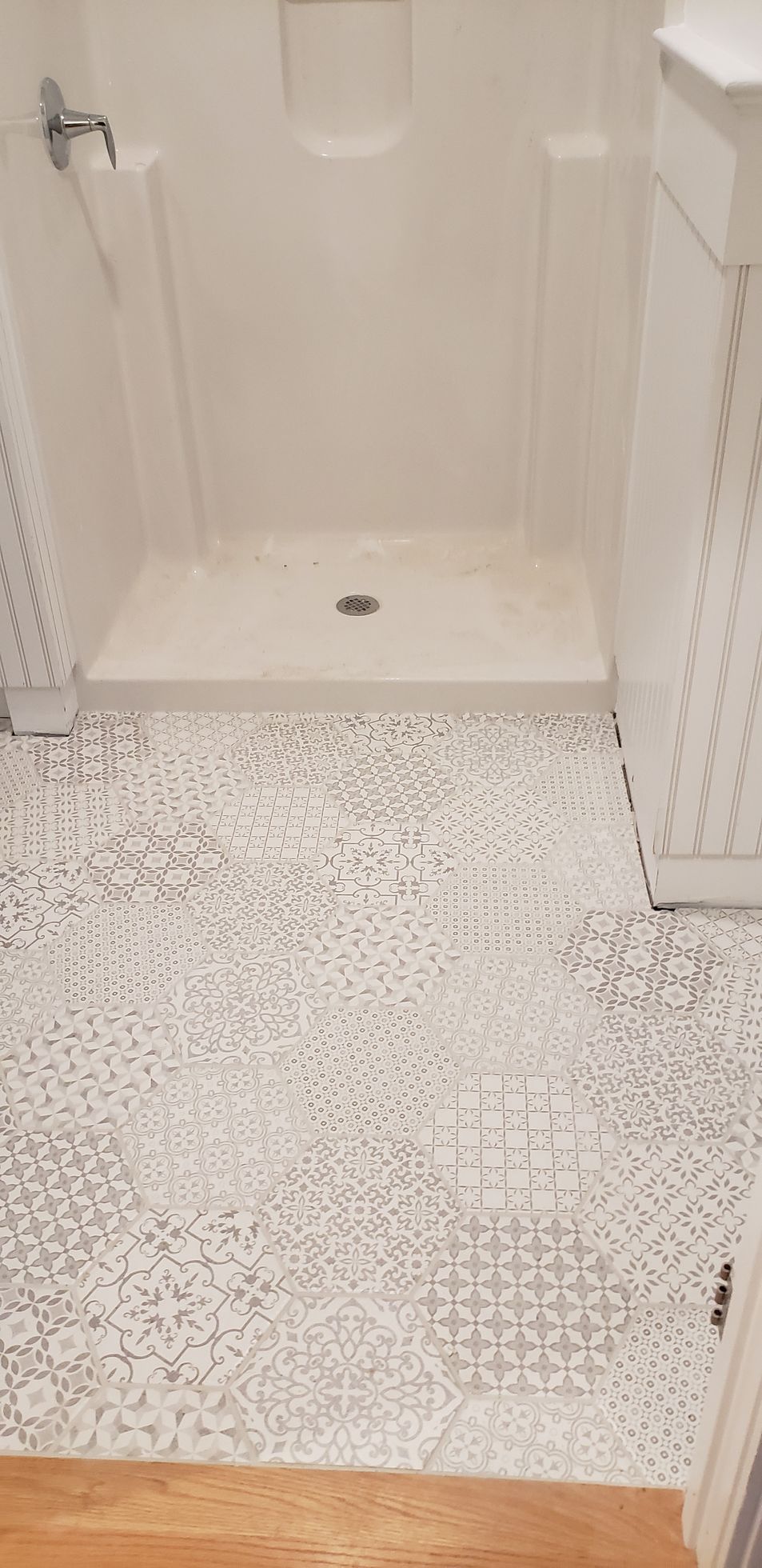 A white shower stall sits on a floor covered in decorative, gray and white patterned hexagonal tiles.