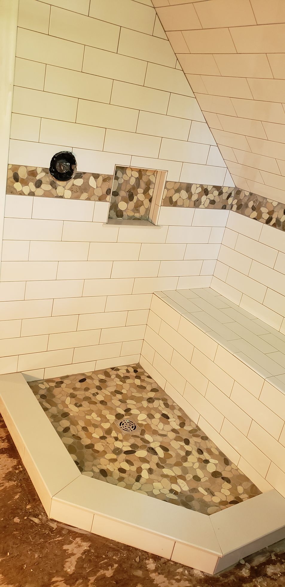 A walk-in shower under construction with white subway tile walls, a pebble-tiled floor, a matching niche, and a stone bench.