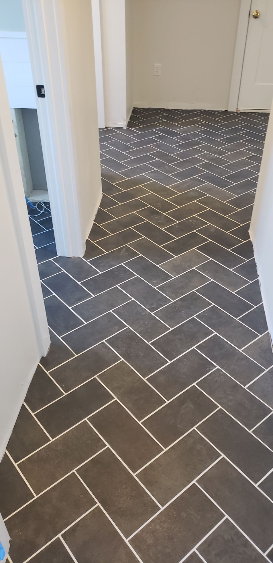 A hallway floor featuring dark gray rectangular tiles laid in a classic herringbone pattern with white grout lines.