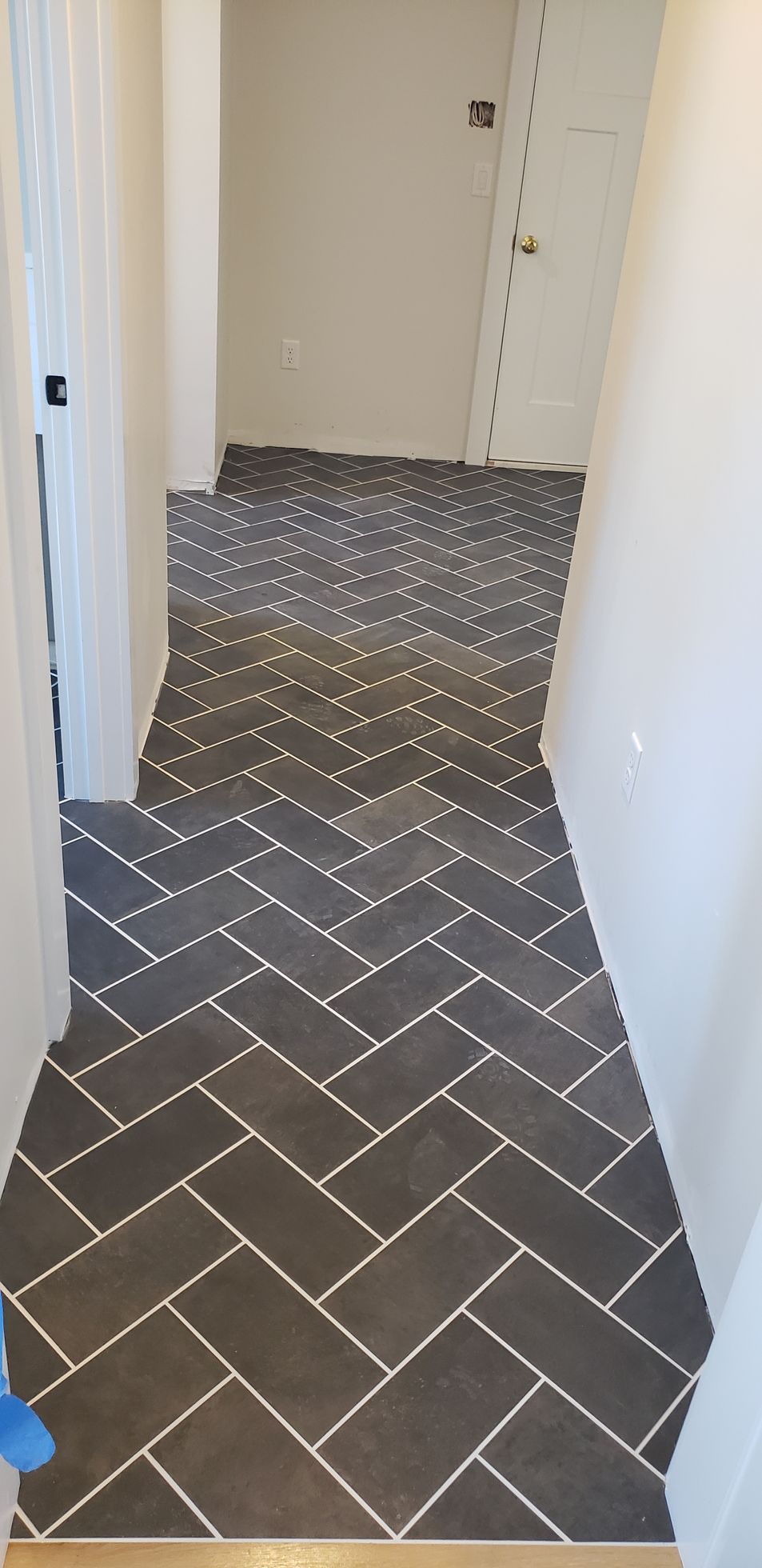 A narrow hallway features dark gray rectangular floor tiles arranged in a herringbone pattern with white grout.