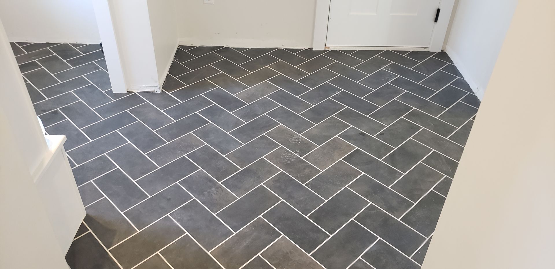 Gray rectangular tiles laid in a herringbone pattern on a floor inside a room with white walls and a white door.