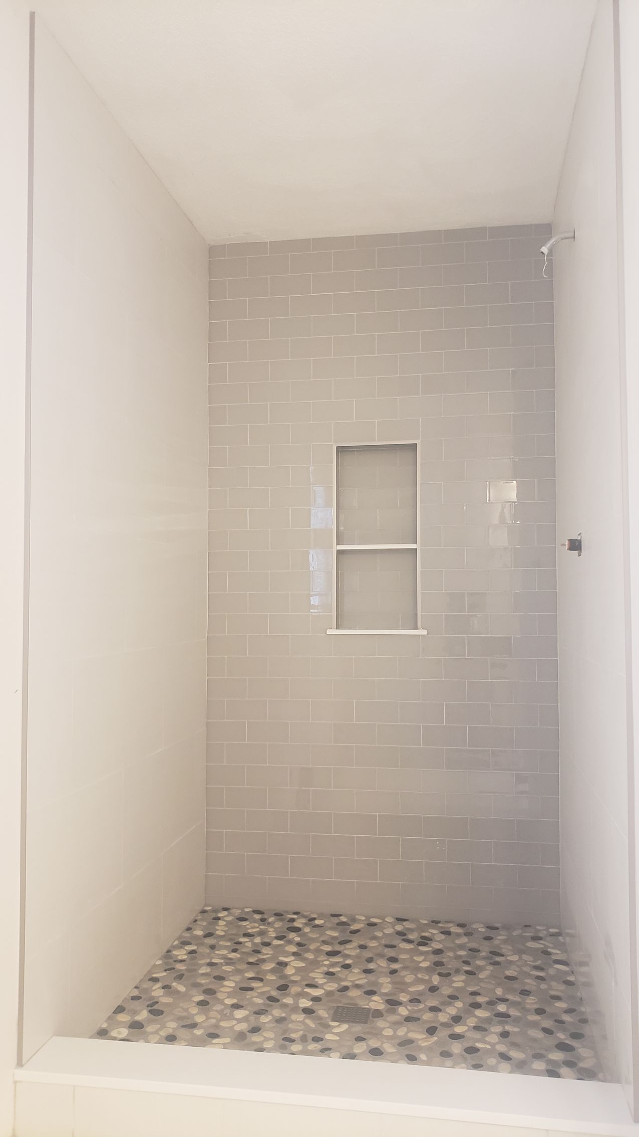 A walk-in shower with light grey subway tile walls, a central built-in niche, and a grey pebble stone floor.